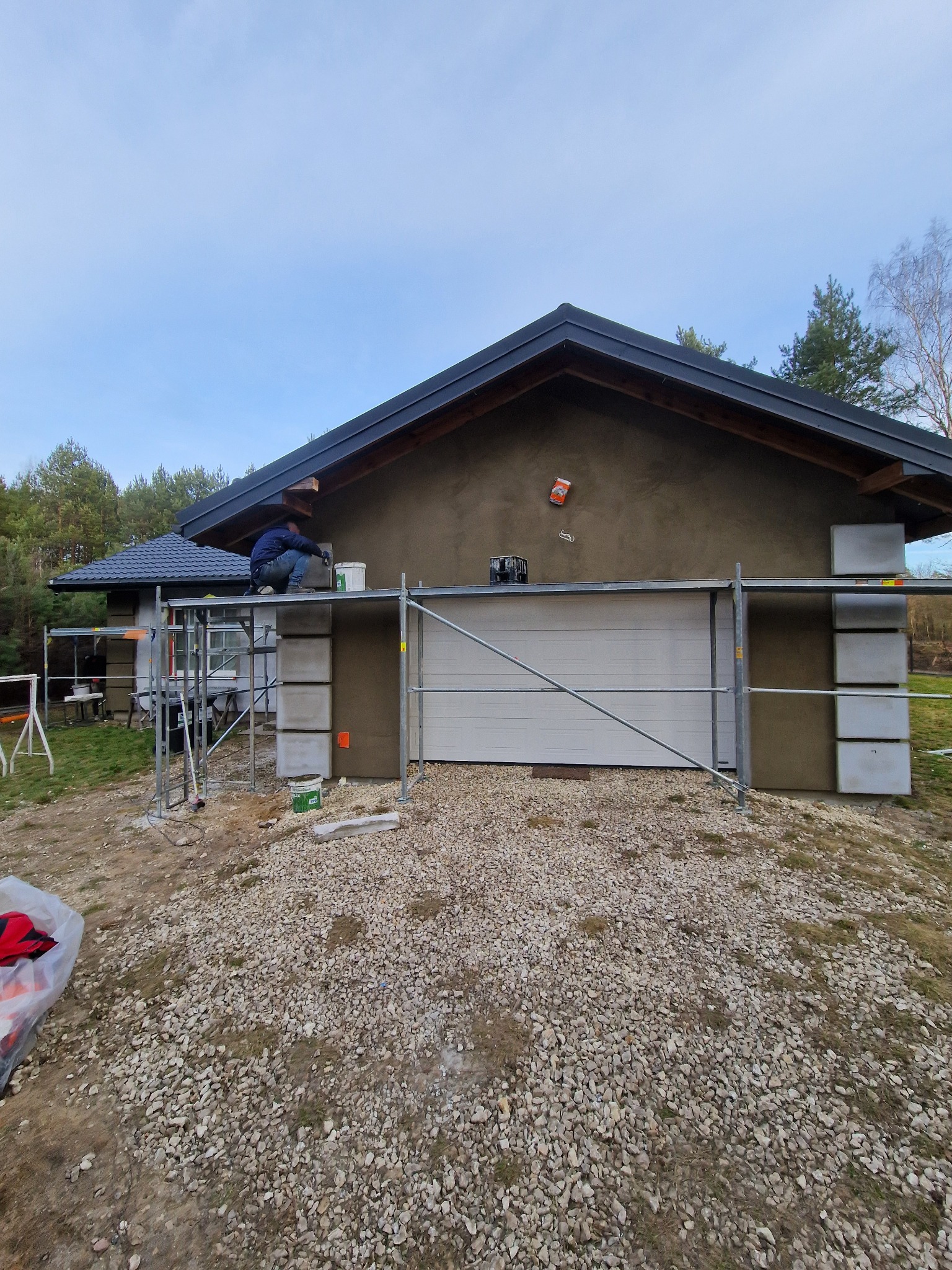 Pracownik na rusztowaniu nakłada warstwę tynku na elewację budynku z garażem, widoczne płyty izolacyjne i narzędzia.