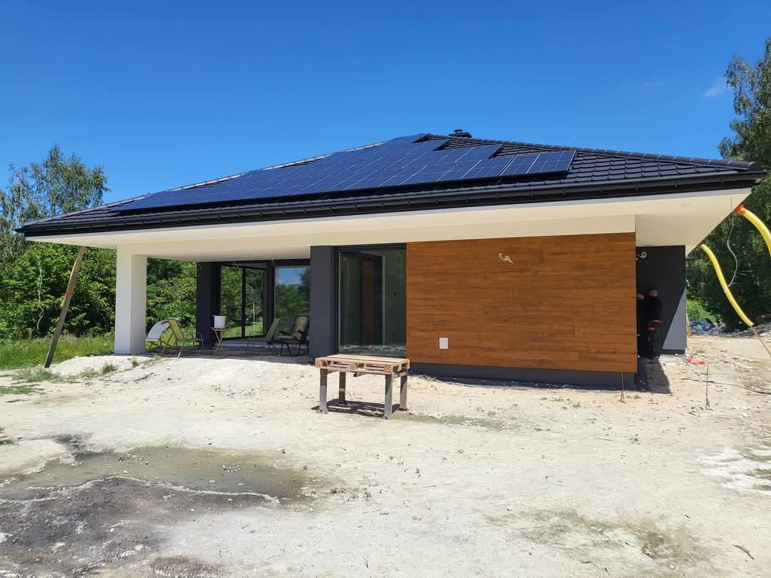 Nowoczesny dom jednorodzinny z panelami słonecznymi na dachu, wykończony drewnianą elewacją i szarymi ścianami, widoczny z zewnątrz na etapie prac wykończeniowych, z piaskiem na podwórku...