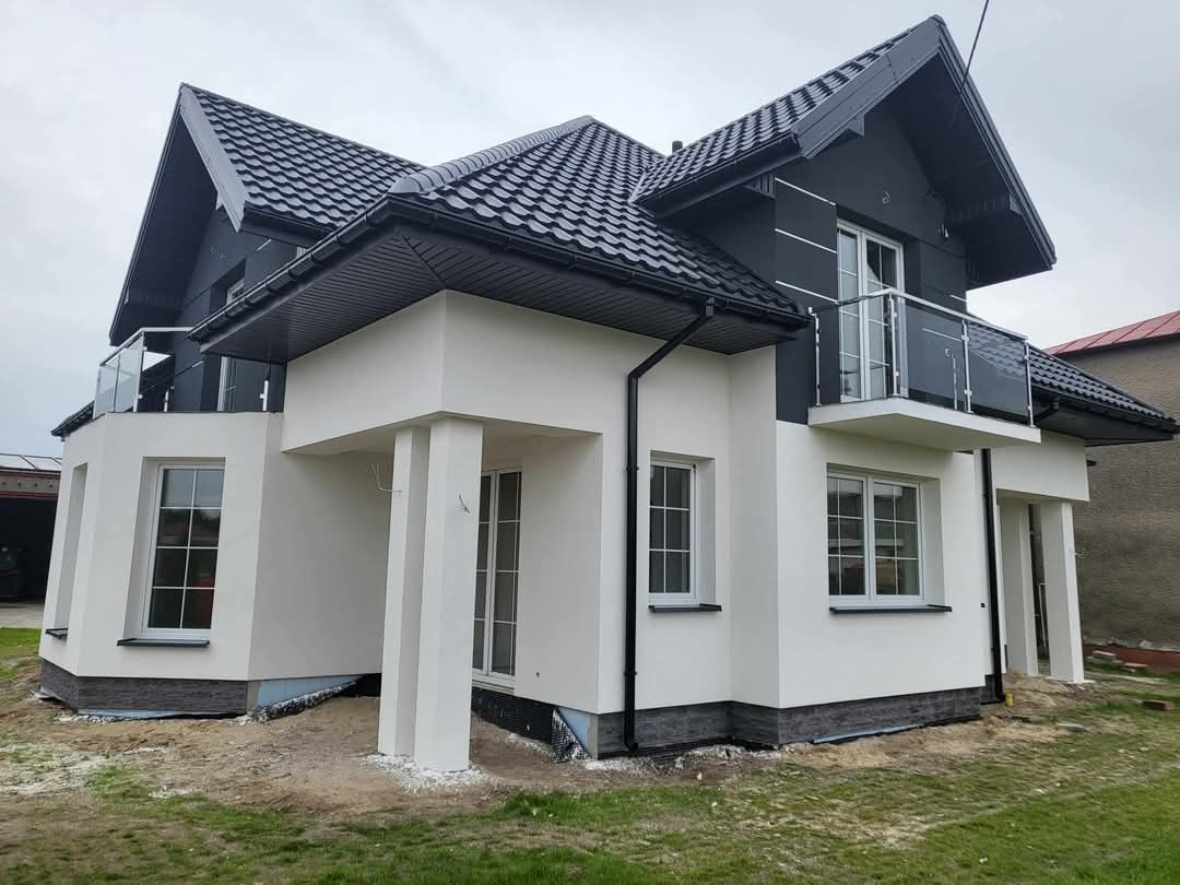 Nowoczesny dom jednorodzinny po remoncie elewacji, z białymi ścianami, ciemnoszarą dachówką i balkonami z szklanymi balustradami, widoczne detale wykończenia.