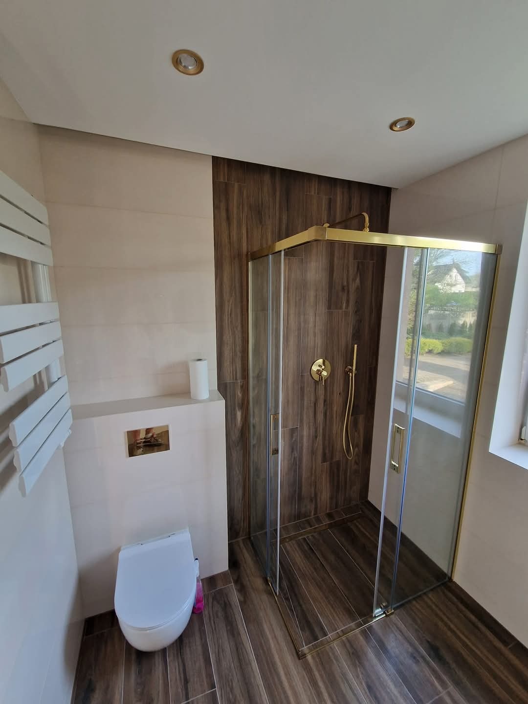 Nowoczesna łazienka z kabiną prysznicową ze złotymi ramami, toaletą podwieszaną i grzejnikiem drabinkowym, wykończona płytkami imitującymi drewno i beżowymi panelami.