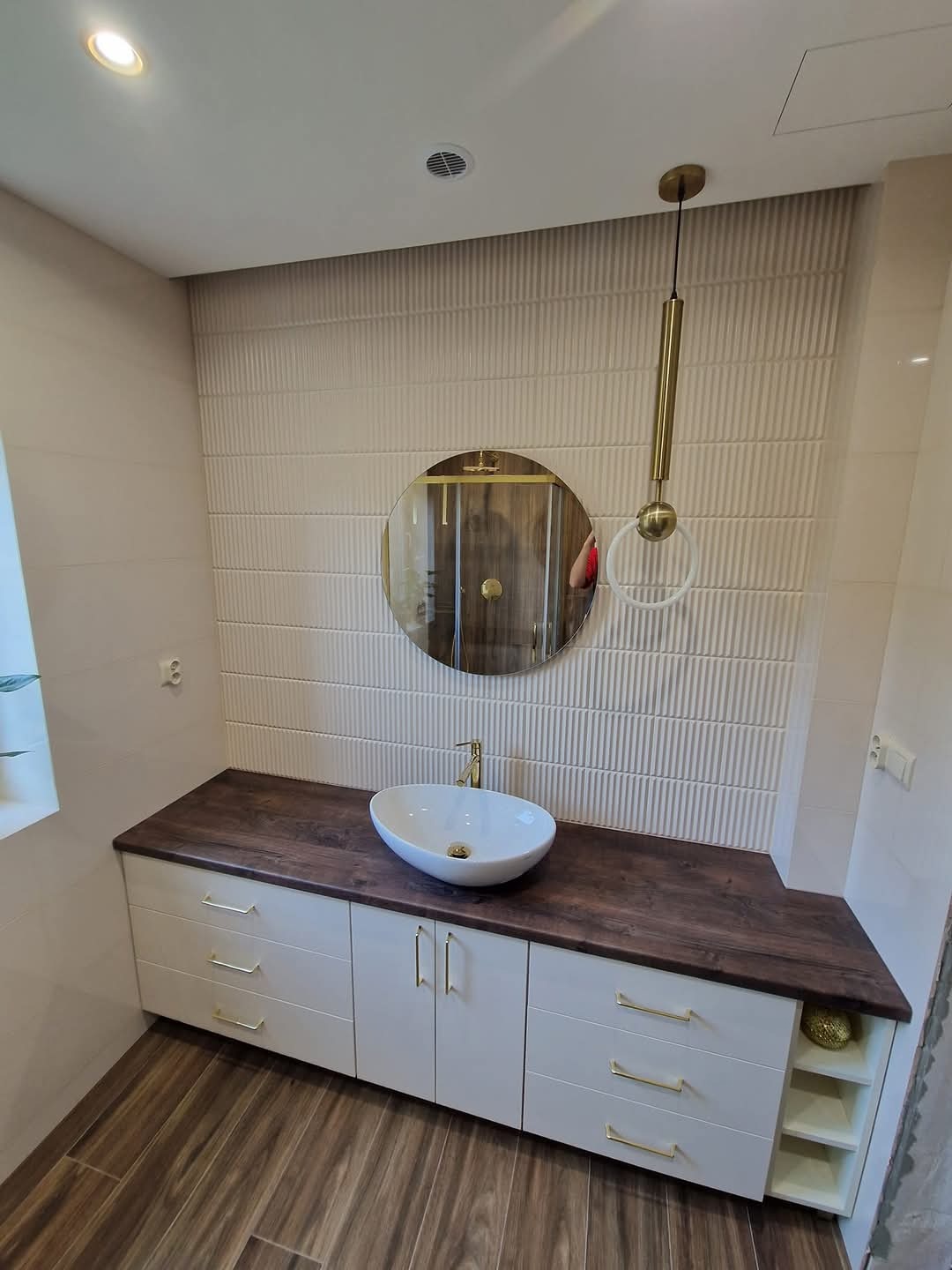 Nowoczesna łazienka z owalną umywalką nablatową na ciemnym drewnianym blacie, białe szafki z złotymi uchwytami, okrągłe lustro i dekoracyjna lampa wisząca na tle ściany z białych, teksturowanych...