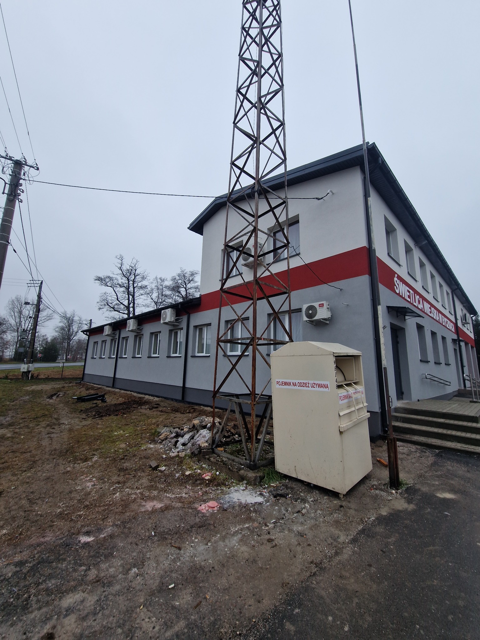 Budynek świetlicy wiejskiej z szarą elewacją i czerwonym pasem, widoczny z zewnątrz, obok metalowej wieży i kontenera na odzież używaną.