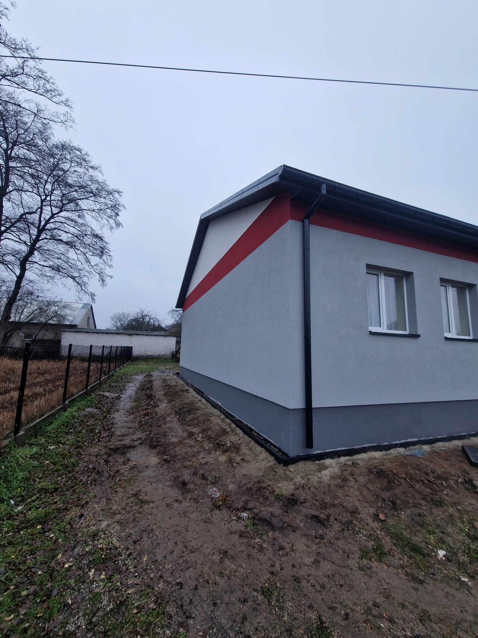 Świeżo otynkowany budynek z szarą elewacją i czerwonym pasem pod dachem, widoczne okna z białymi ramami, czarna rynna, obok błotnista ścieżka i ogrodzenie.