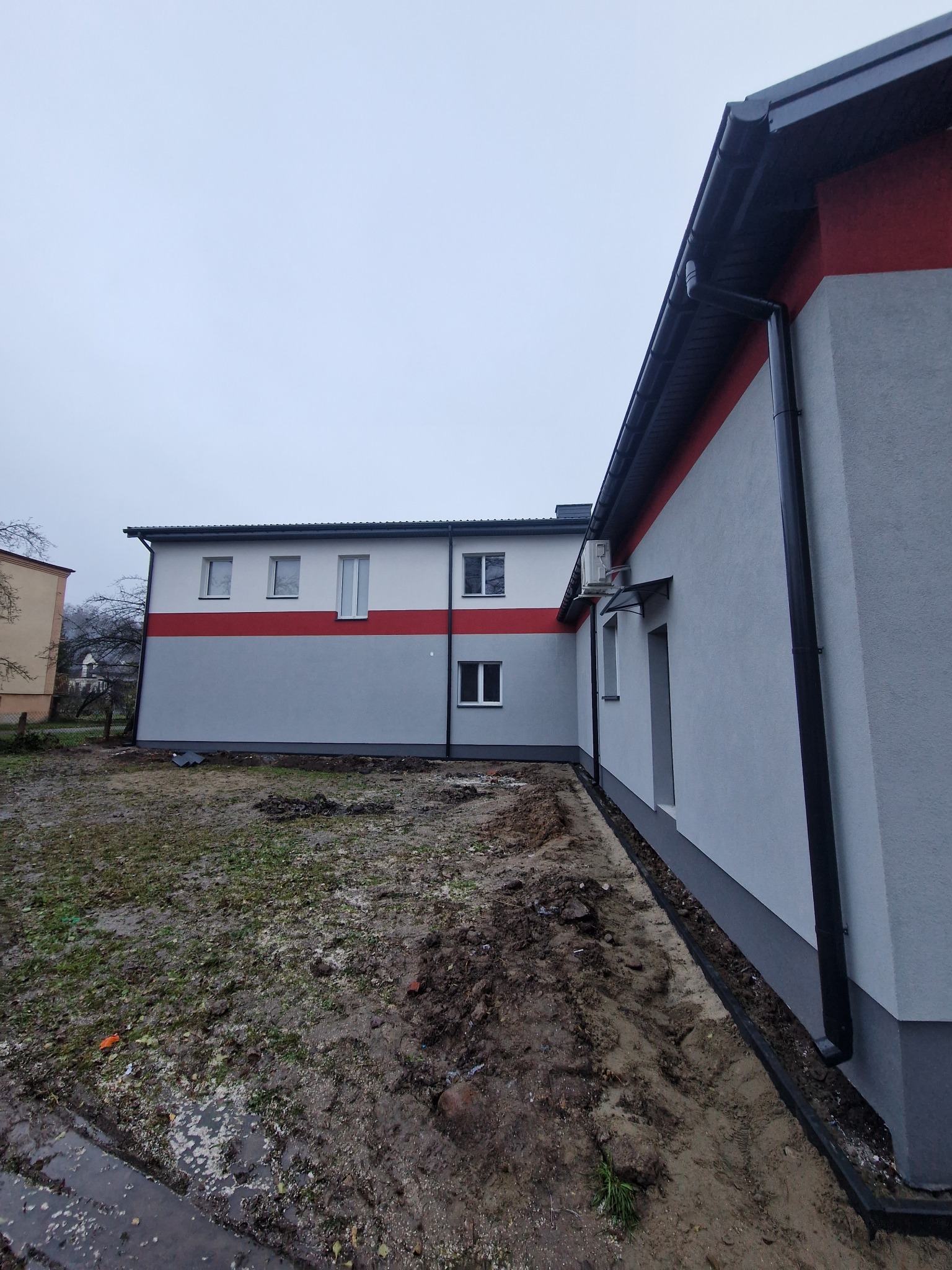 Budynek po remoncie elewacji z widocznym ociepleniem i nowym orynnowaniem, w tle drugi budynek, teren wokół budynku częściowo rozkopany.