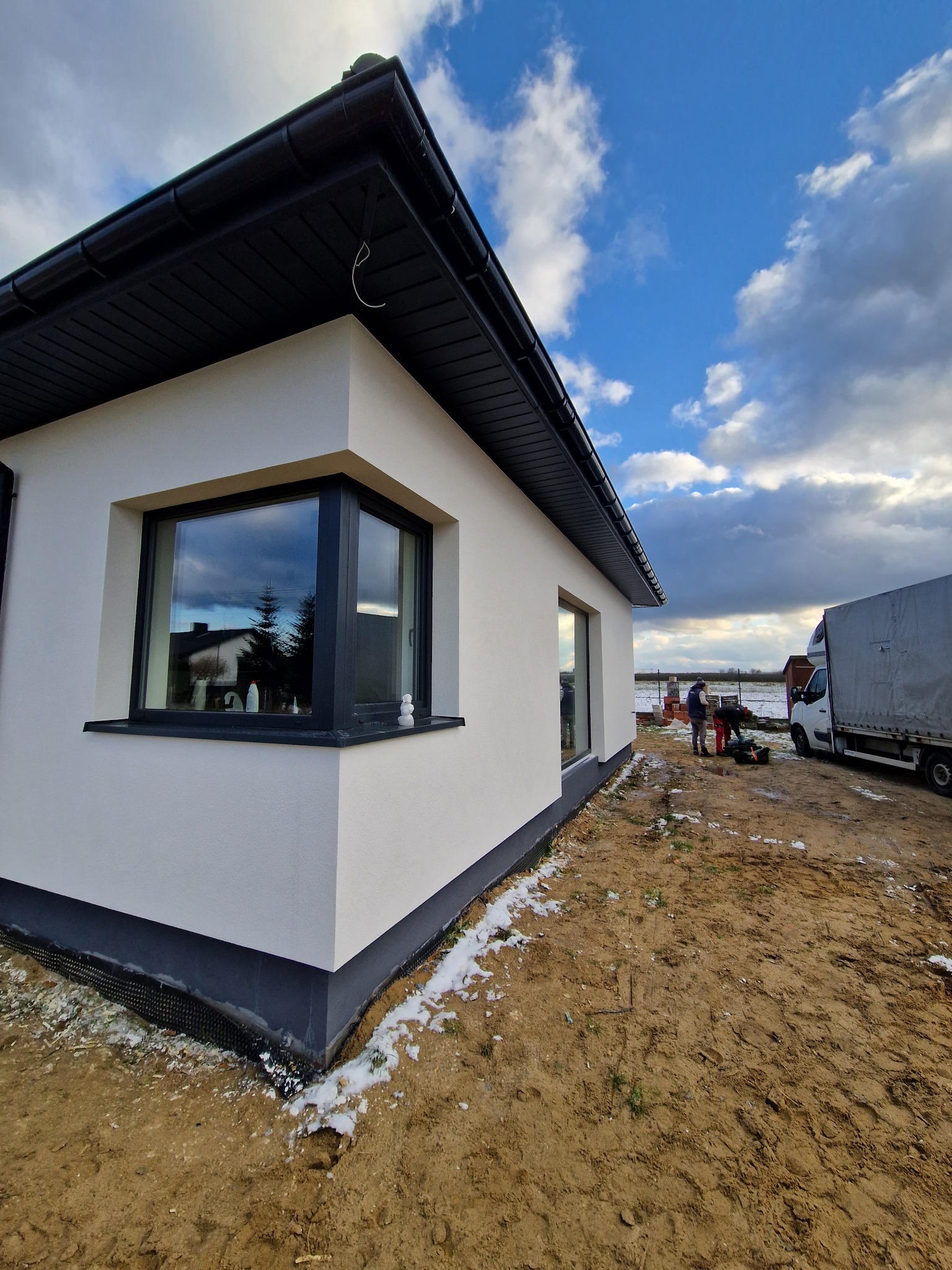 Nowoczesny, parterowy dom z ocieploną elewacją w kolorze białym, z czarną stolarką okienną i dachem, widoczne prace wykończeniowe na zewnątrz budynku, częściowo ośnieżony teren.