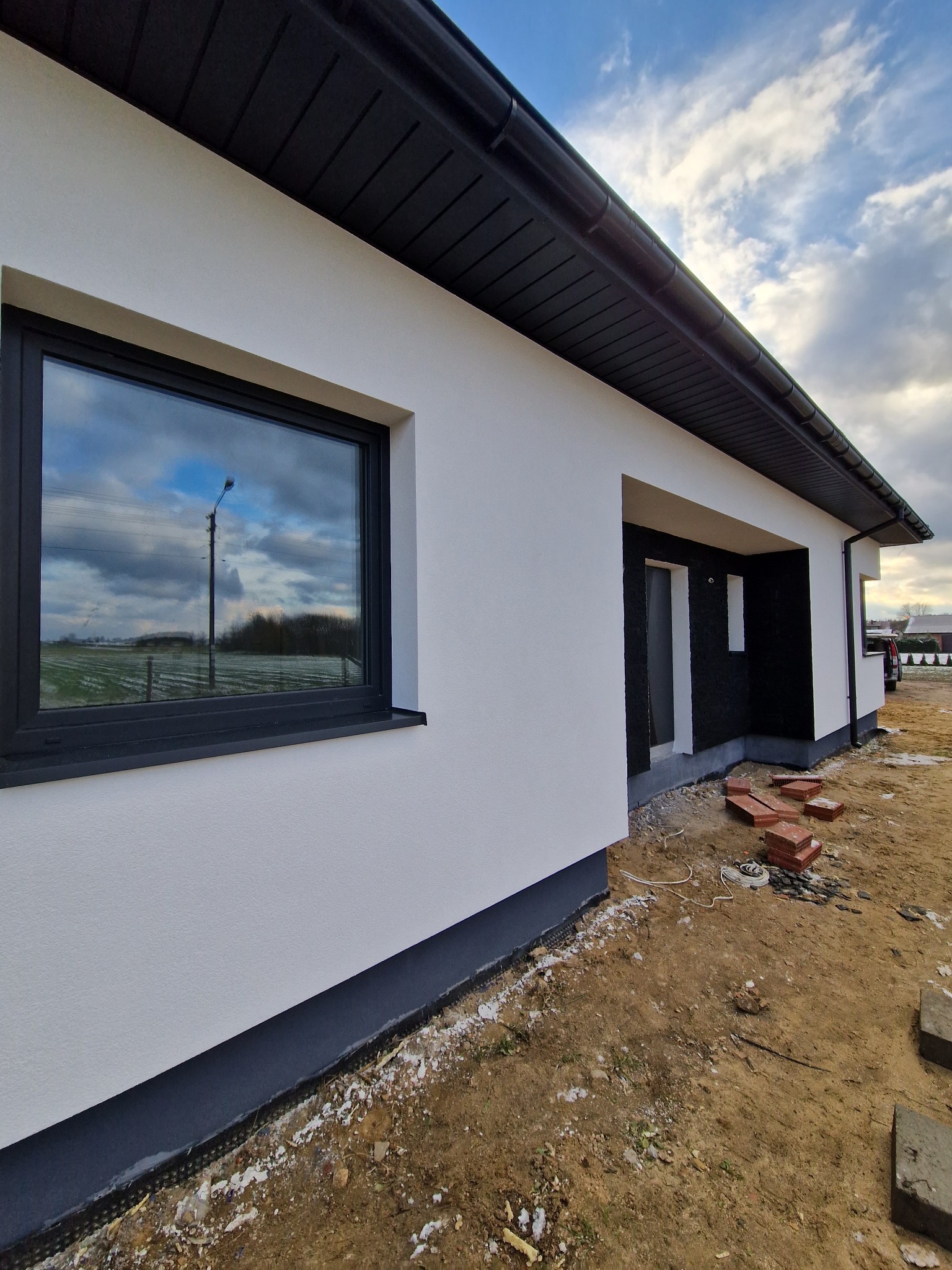 Nowo otynkowana ściana domu jednorodzinnego z czarną stolarką okienną i wykończeniem dachu, widoczne cegły na ziemi i przygotowane wejście do budynku.