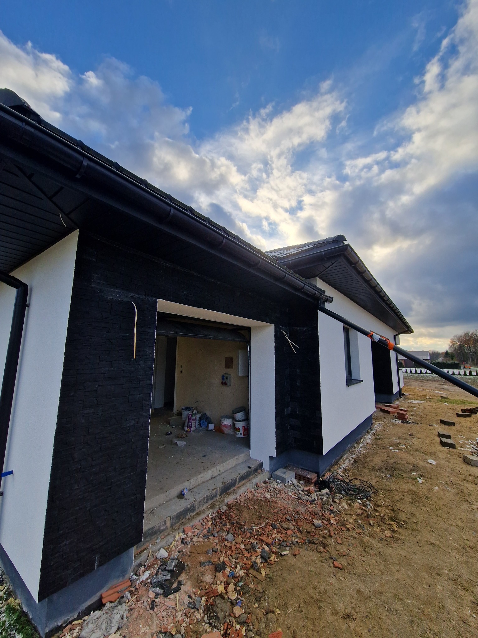 Nowoczesny dom jednorodzinny z czarno-białą elewacją, widoczne fragmenty ocieplenia i przygotowania do prac wykończeniowych, niezagospodarowany teren wokół budynku.