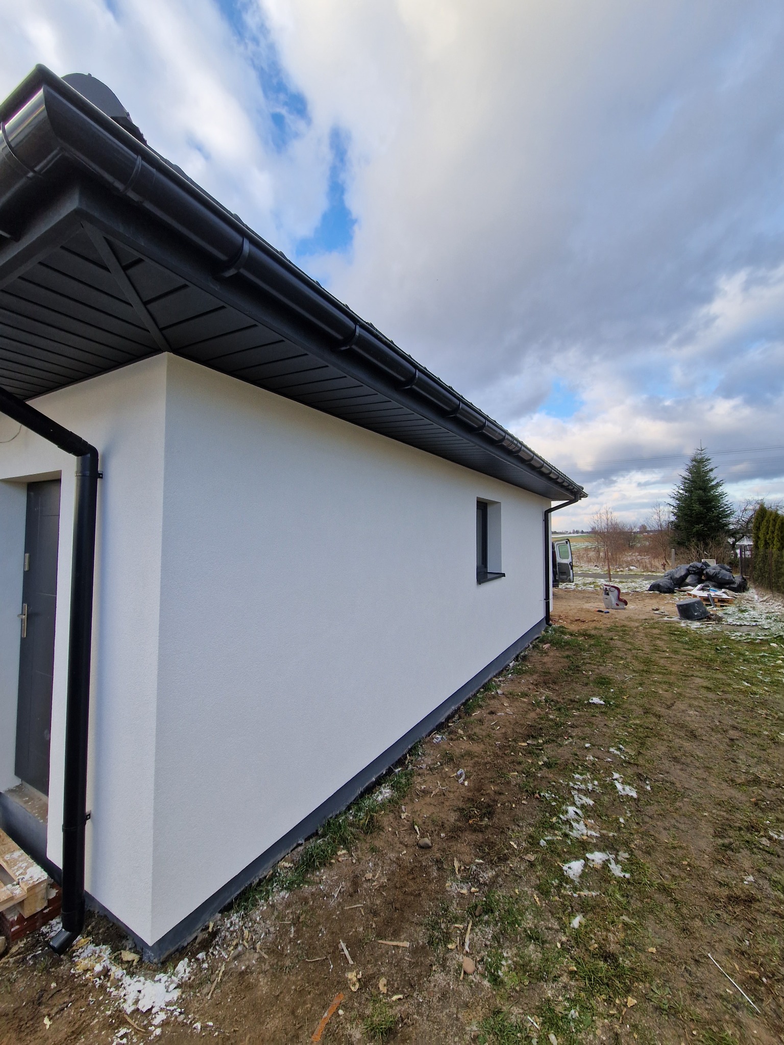 Boczny widok budynku z białą elewacją, czarnymi rynnami i niewielkim oknem. Na ziemi resztki śniegu, w tle widoczne drzewa i pochmurne niebo.