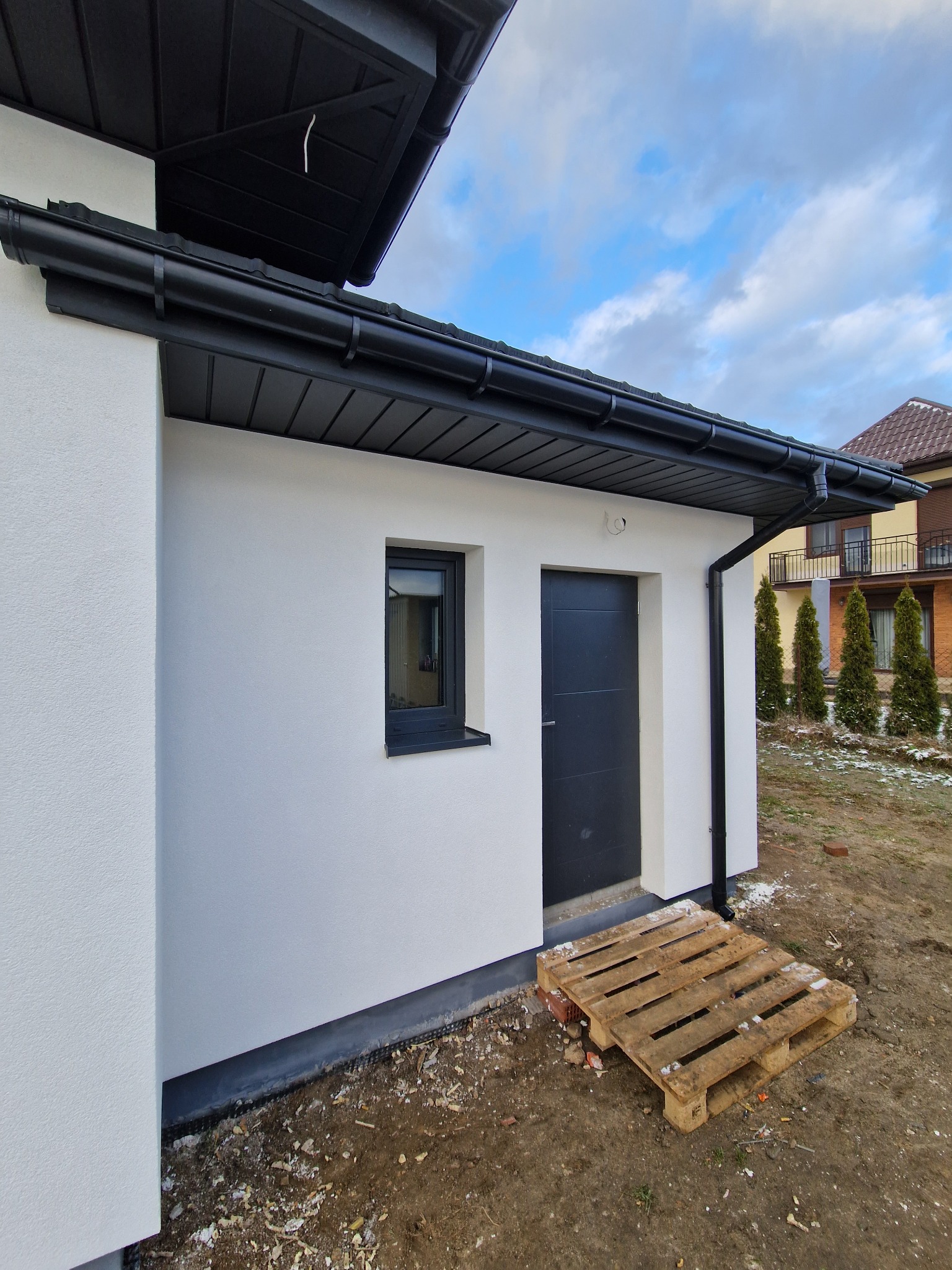Biała elewacja budynku z czarną stolarką okienną i drzwiami, czarne orynnowanie, paleta drewniana przed wejściem, widok na sąsiedni dom i kilka tuj