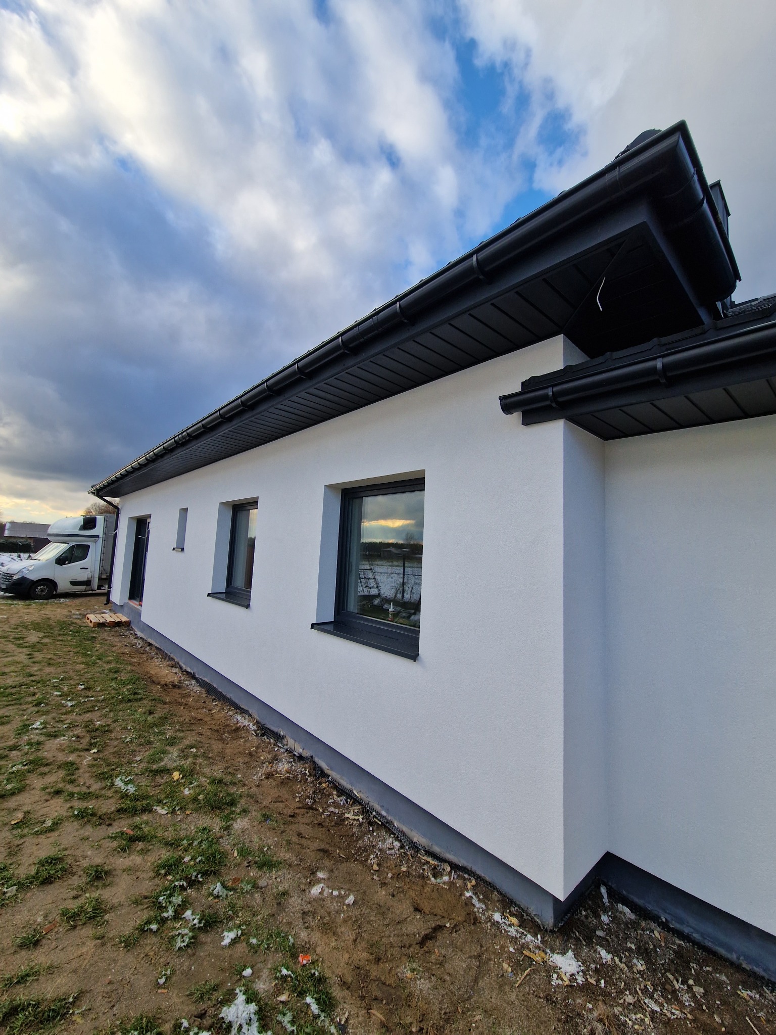 Boczny widok nowo otynkowanego, jednopiętrowego domu z szarymi oknami i czarnymi rynnami, z widoczną izolacją fundamentów i częściowo zagospodarowanym terenem wokół.