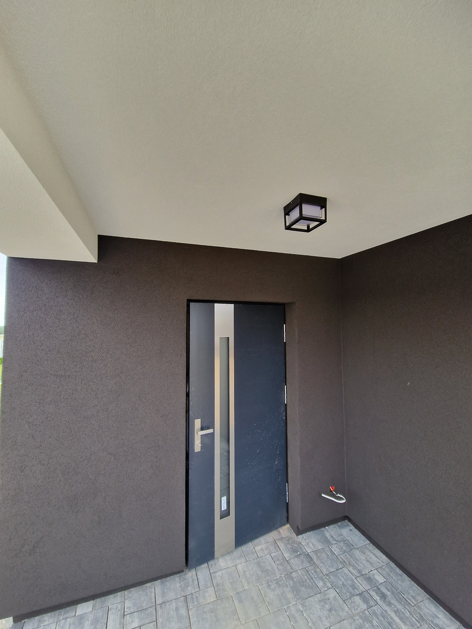 Nowoczesne wejście do domu z ciemnoszarą elewacją o wyraźnej fakturze, stalowe drzwi z przeszkleniem, kostka brukowa na podłodze i minimalistyczna lampa sufitowa.