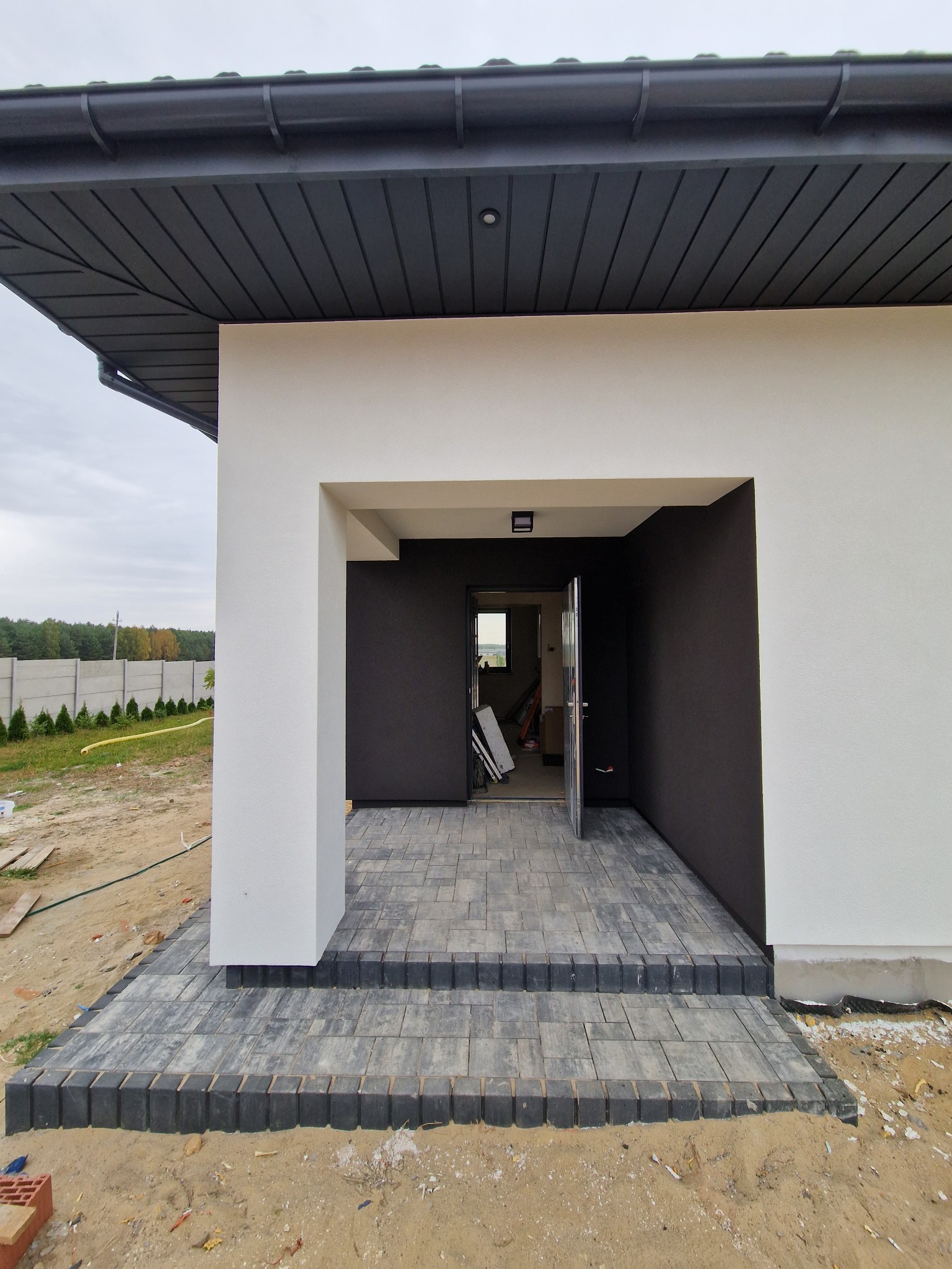 Nowoczesny dom z białymi ścianami, czarnym dachem i wejściem wyłożonym szarą kostką brukową z czarnymi obrzeżami, częściowo otwarte drzwi wejściowe.