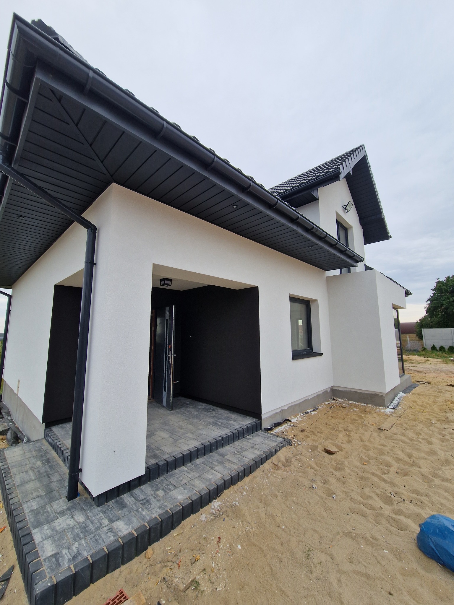 Nowoczesny dom jednorodzinny z białymi ścianami i czarnym dachem oraz stolarką okienną, wykończony kostką brukową przed wejściem, w trakcie prac wykończeniowych terenu.