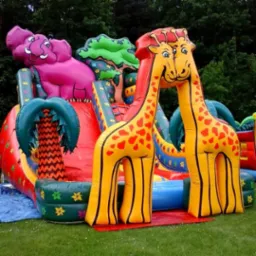 Kolorowy, dmuchany plac zabaw z motywem dżungli, zawierający zjeżdżalnię, dmuchanego słonia i dwie dmuchane żyrafy złączone szyjami, ustawiony na trawniku.