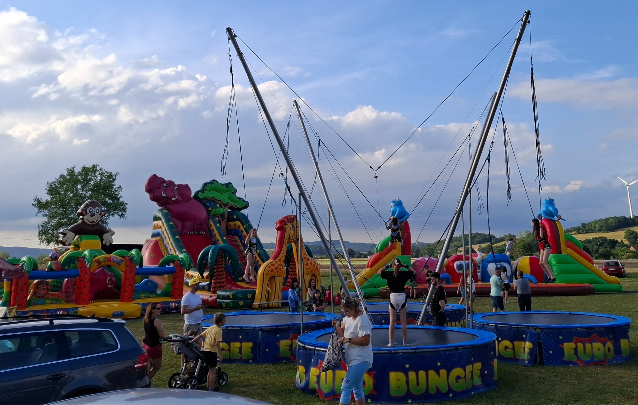 Rozległy plac zabaw na świeżym powietrzu z dmuchanymi zamkami w kształcie zwierząt i stanowiskami do skoków na bungee, w tle widać ludzi i krajobraz z turbiną wiatrową.