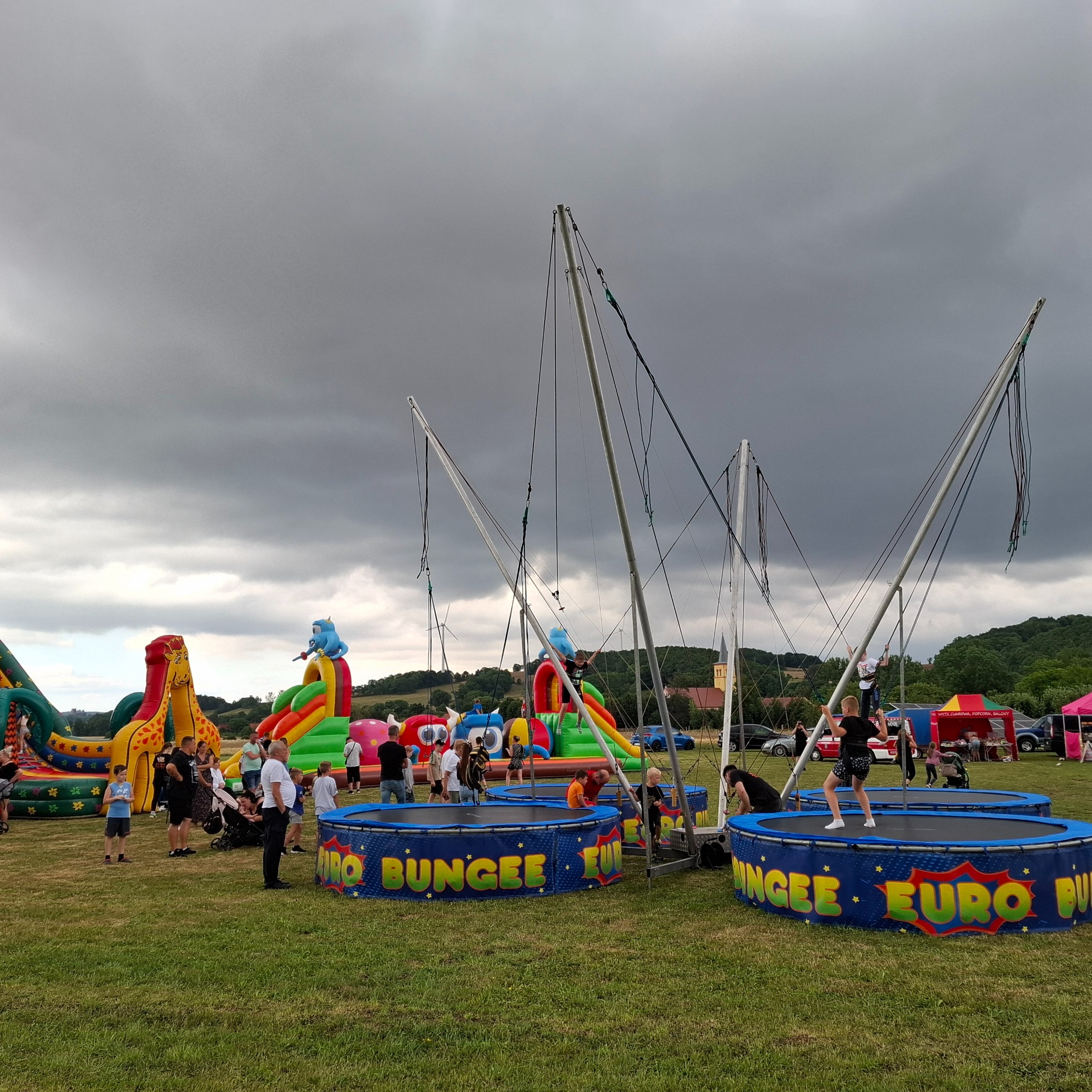 Dziecko skacze na trampolinie bungee na tle dmuchanych zamków i pochmurnego nieba podczas plenerowej imprezy.