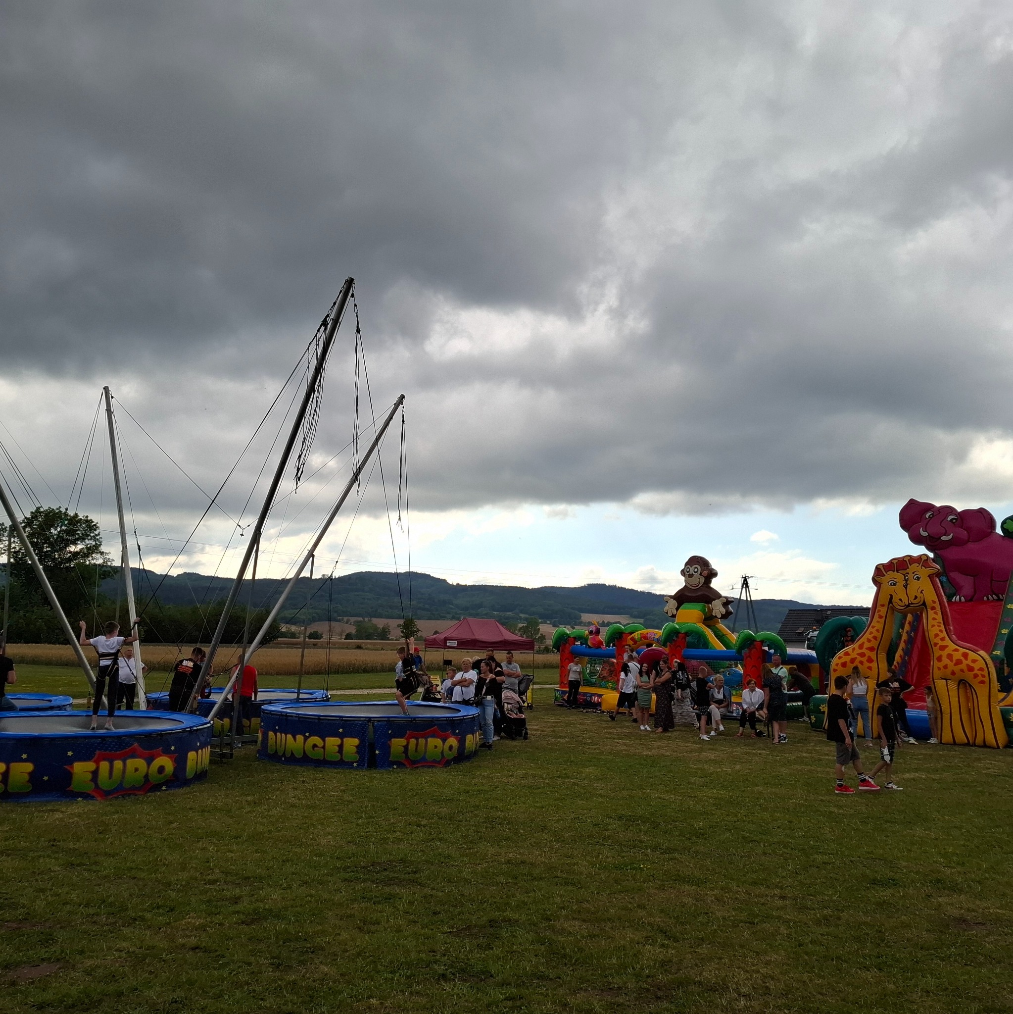 Plenerowa impreza z dmuchanymi atrakcjami: trampoliny bungee z osobami skaczącymi, kolorowy dmuchany plac zabaw z motywami zwierząt, oraz zgromadzeni ludzie na trawiastym terenie pod pochmurnym...