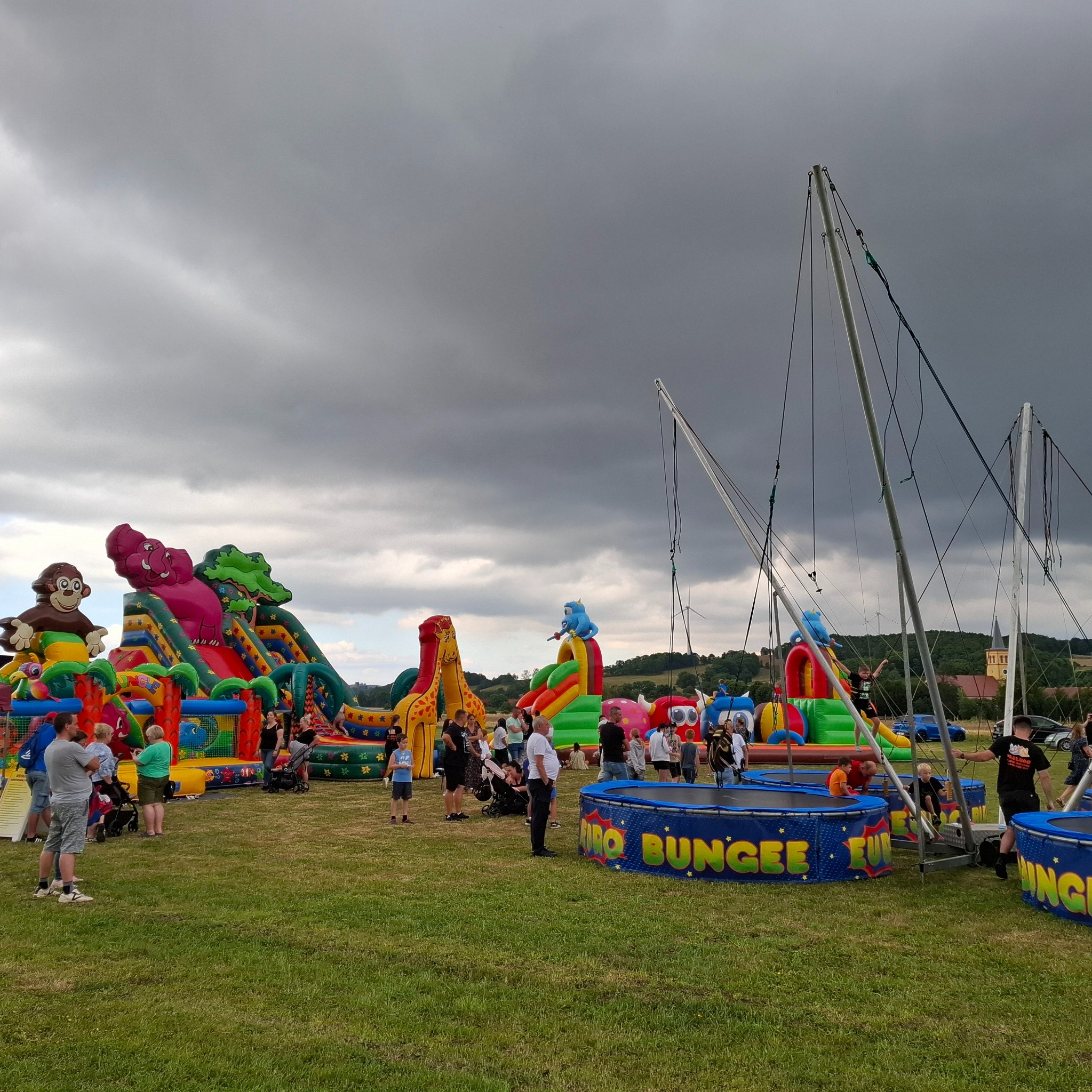 Kolorowe dmuchane zamki ze zwierzętami i trampoliny bungee na trawie w Grochowej, z widocznymi ludźmi i pochmurnym niebem w tle.