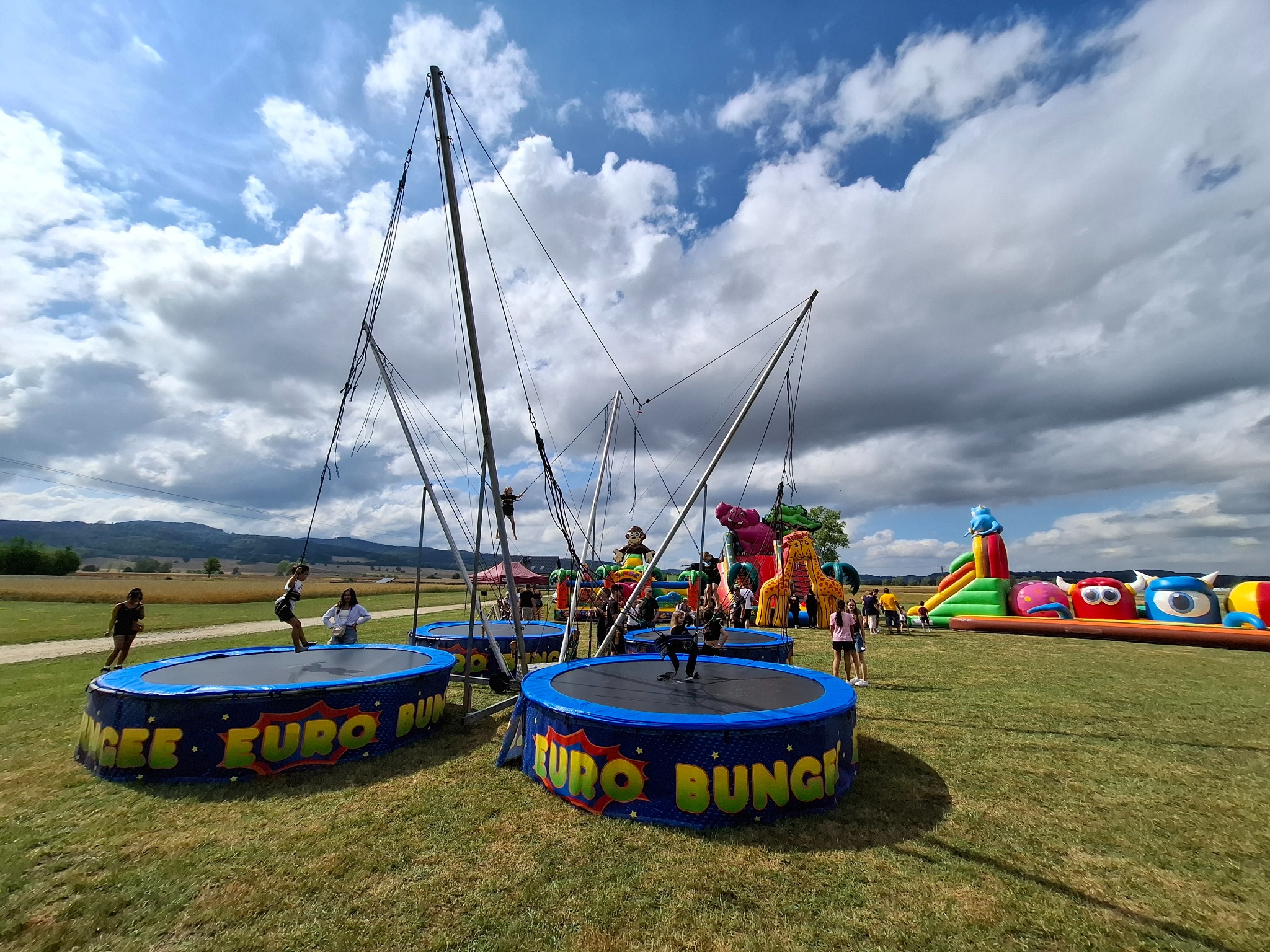 Cztery trampoliny bungee EURO BUNGEE na zielonej trawie, w tle dmuchany plac zabaw i pochmurne niebo.