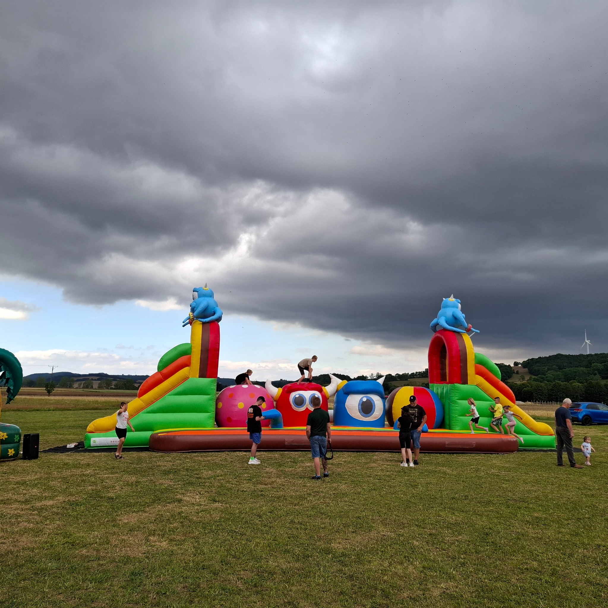 Dmuchany plac zabaw z motywem potworów na otwartej przestrzeni, dzieci bawią się pod pochmurnym niebem, w tle widoczny wiatrak.