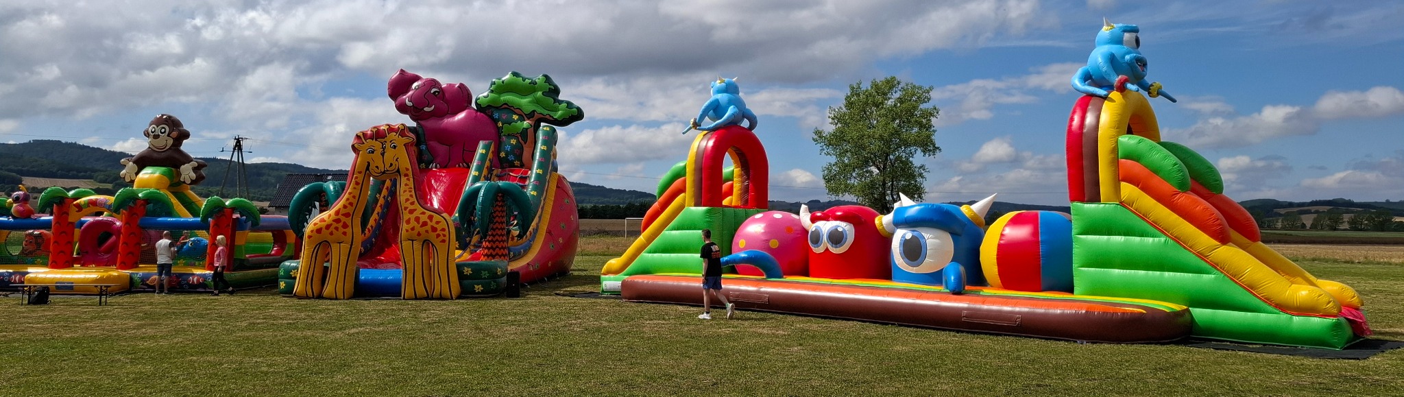 Panoramiczne ujęcie rozległego placu zabaw z dmuchanymi konstrukcjami w kształcie zwierząt i postaci z bajek, ustawionymi na zielonej trawie pod błękitnym niebem z chmurami, z widocznymi osobami...