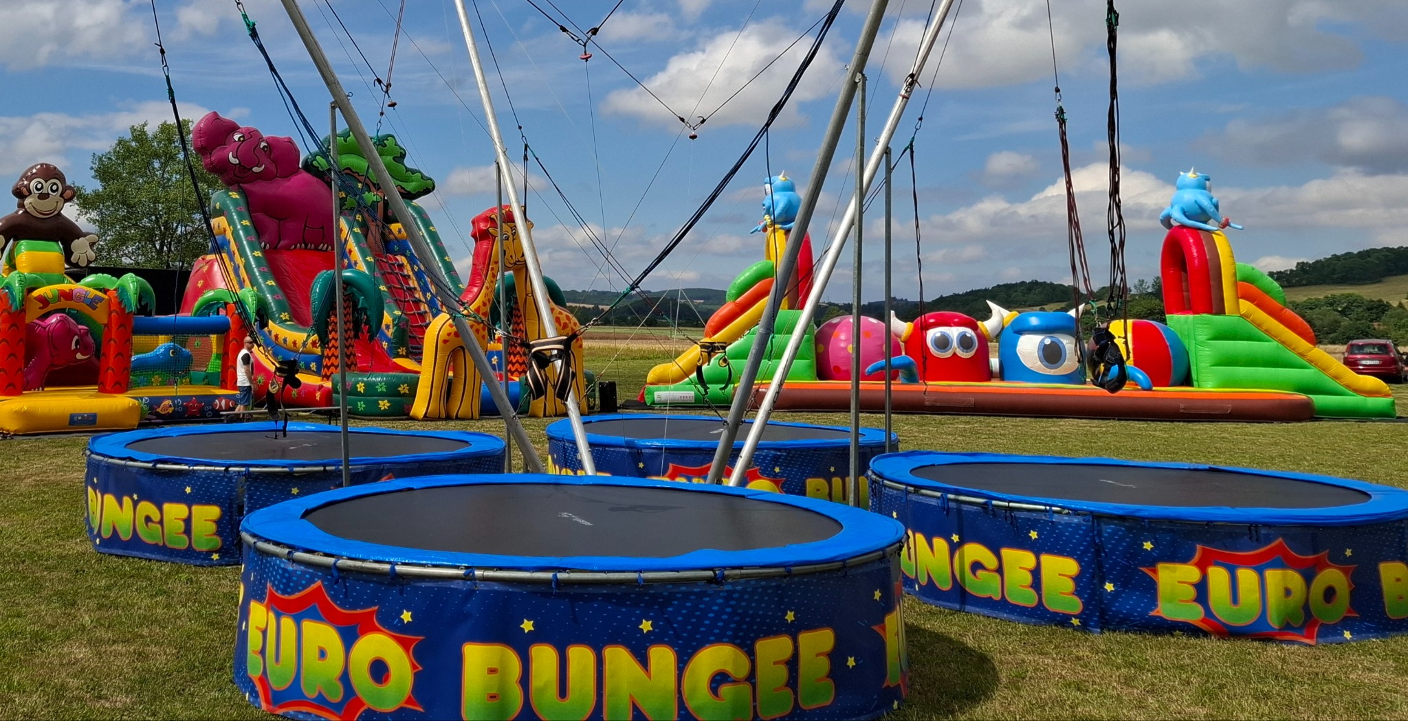 Kompleks rekreacyjny na trawie: kolorowe dmuchane zamki ze zjeżdżalniami w kształcie zwierząt i trampoliny bungee z niebieskimi obudowami z napisem EURO BUNGEE, rozstawione na imprezie plenerowej.