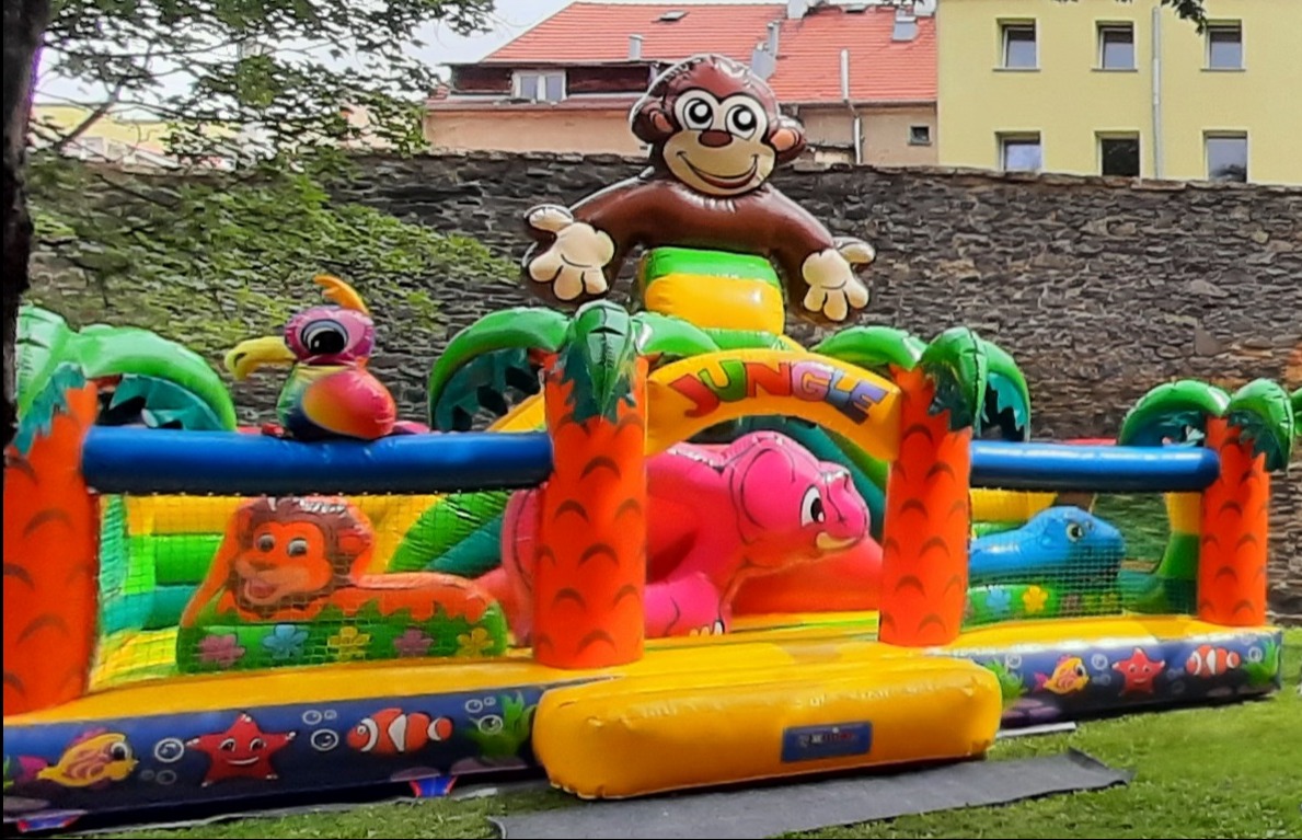 Kolorowy, dmuchany plac zabaw w motywie dżungli z małpą na szczycie, lwem, słoniem i krokodylem, ustawiony na trawie przed kamiennym murem i budynkiem.