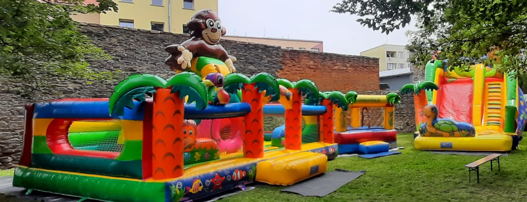 Kompleksowy, kolorowy park rozrywki dmuchanej, z motywami dżungli i małpą na szczycie, ustawiony na trawniku przed murem z kamienia i cegły.