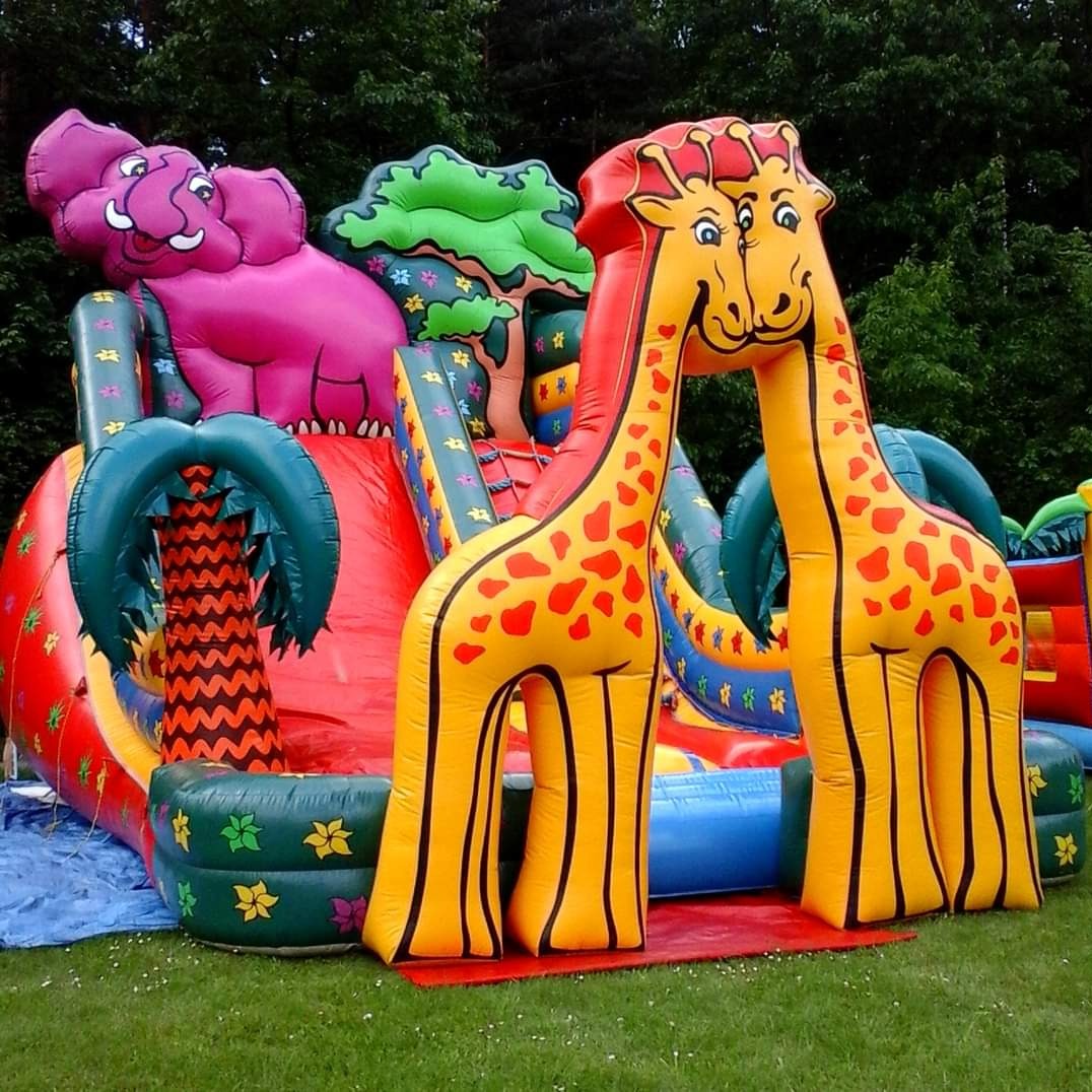 Kolorowy, dmuchany plac zabaw z motywem dżungli, zawierający zjeżdżalnię, dmuchanego słonia i dwie dmuchane żyrafy złączone szyjami, ustawiony na trawniku.