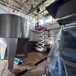 MuchiClean - Wnętrze przemysłowe z dużym stalowym zbiornikiem, posadzka z płytek, podnośnik nożycowy z pracownikiem przy suficie i maszyna zakryta folią ochronną.