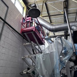 MuchiClean - Podnośnik nożycowy z pracownikiem w koszu na tle przemysłowego wnętrza z instalacjami i ścianami z płytek, prawdopodobnie podczas prac porządkowych lub konserwacyjnych.