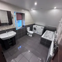 Wnętrze łazienki na poddaszu z umywalką, toaletą, wanną i pralko-suszarką, wykończone płytkami w odcieniach szarości i bieli.