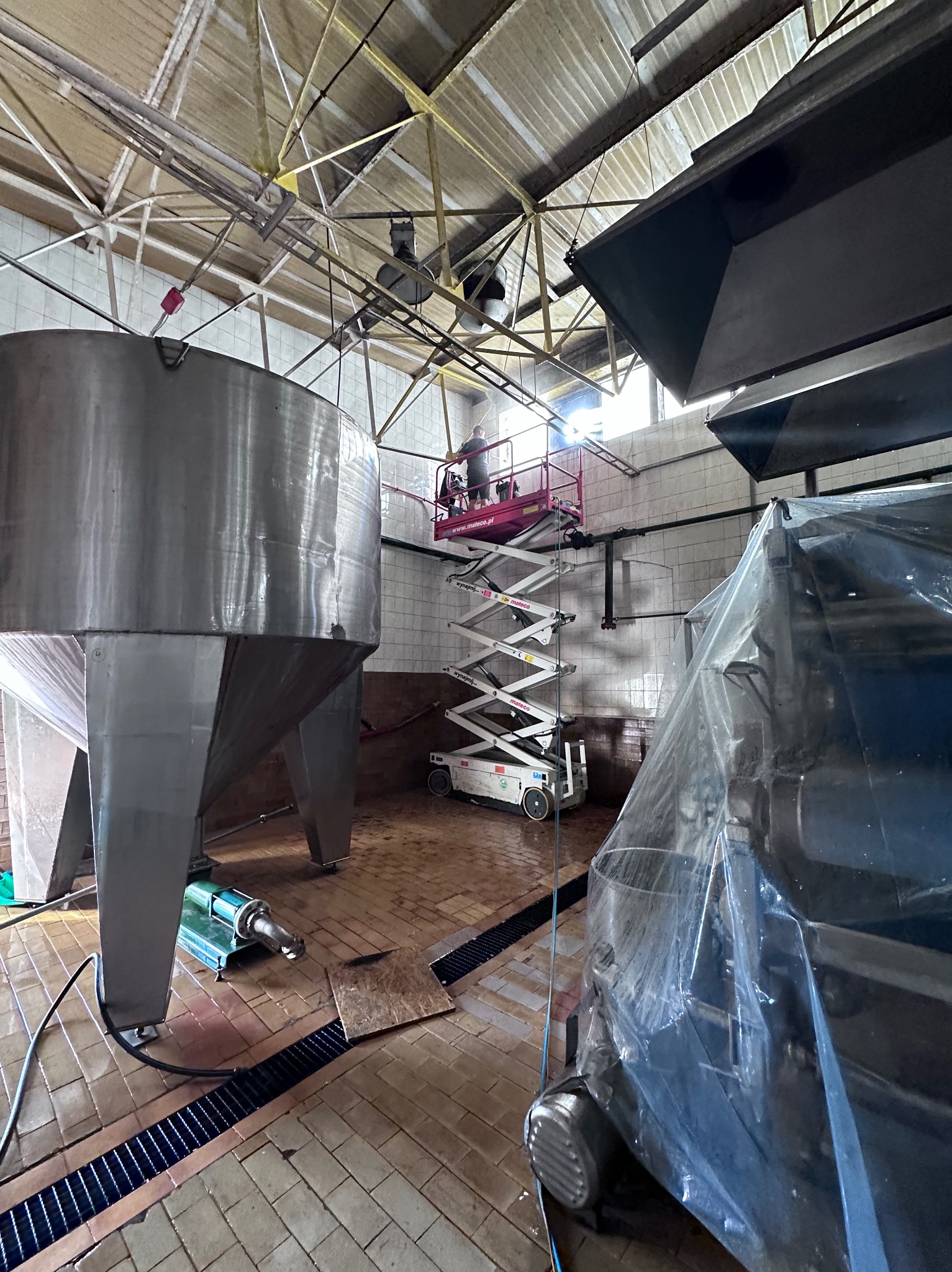 Wnętrze przemysłowe z dużym stalowym zbiornikiem, posadzka z płytek, podnośnik nożycowy z pracownikiem przy suficie i maszyna zakryta folią ochronną.