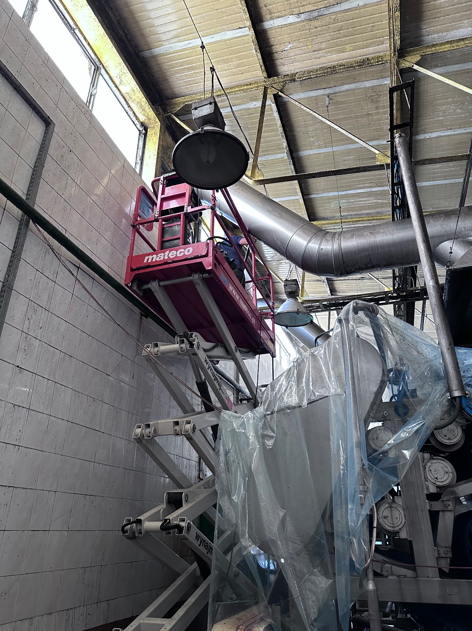 Podnośnik nożycowy z pracownikiem w koszu na tle przemysłowego wnętrza z instalacjami i ścianami z płytek, prawdopodobnie podczas prac porządkowych lub konserwacyjnych.