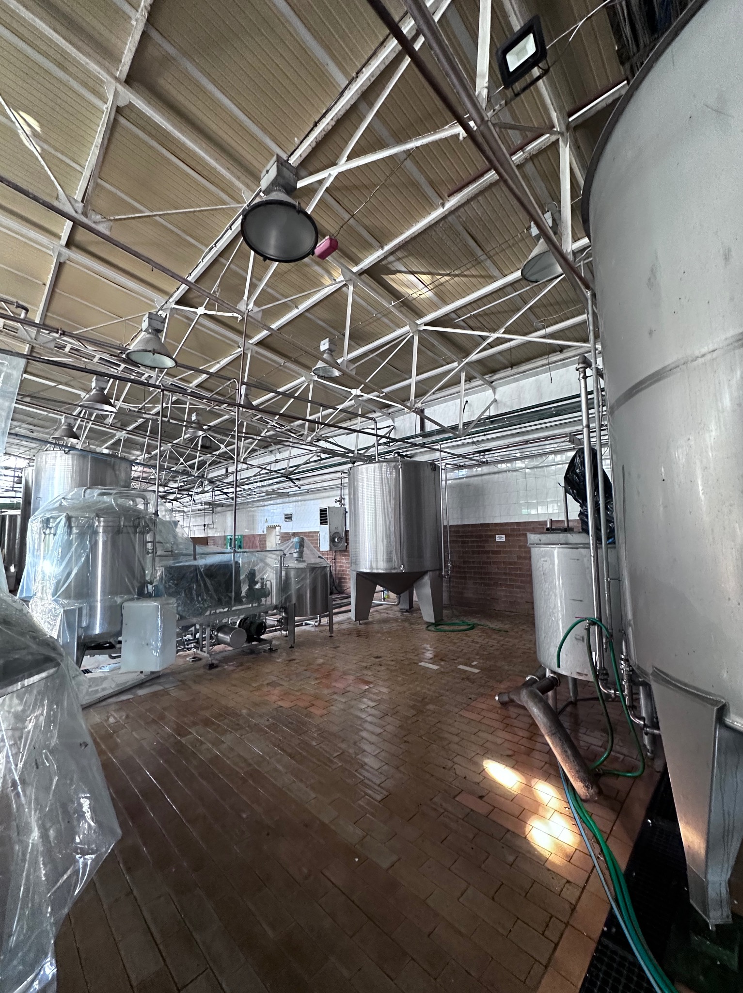 Wnętrze hali produkcyjnej z dużymi, metalowymi zbiornikami, niektóre z nich owinięte folią ochronną, oświetlone lampami przemysłowymi, z posadzką wyłożoną cegłą.