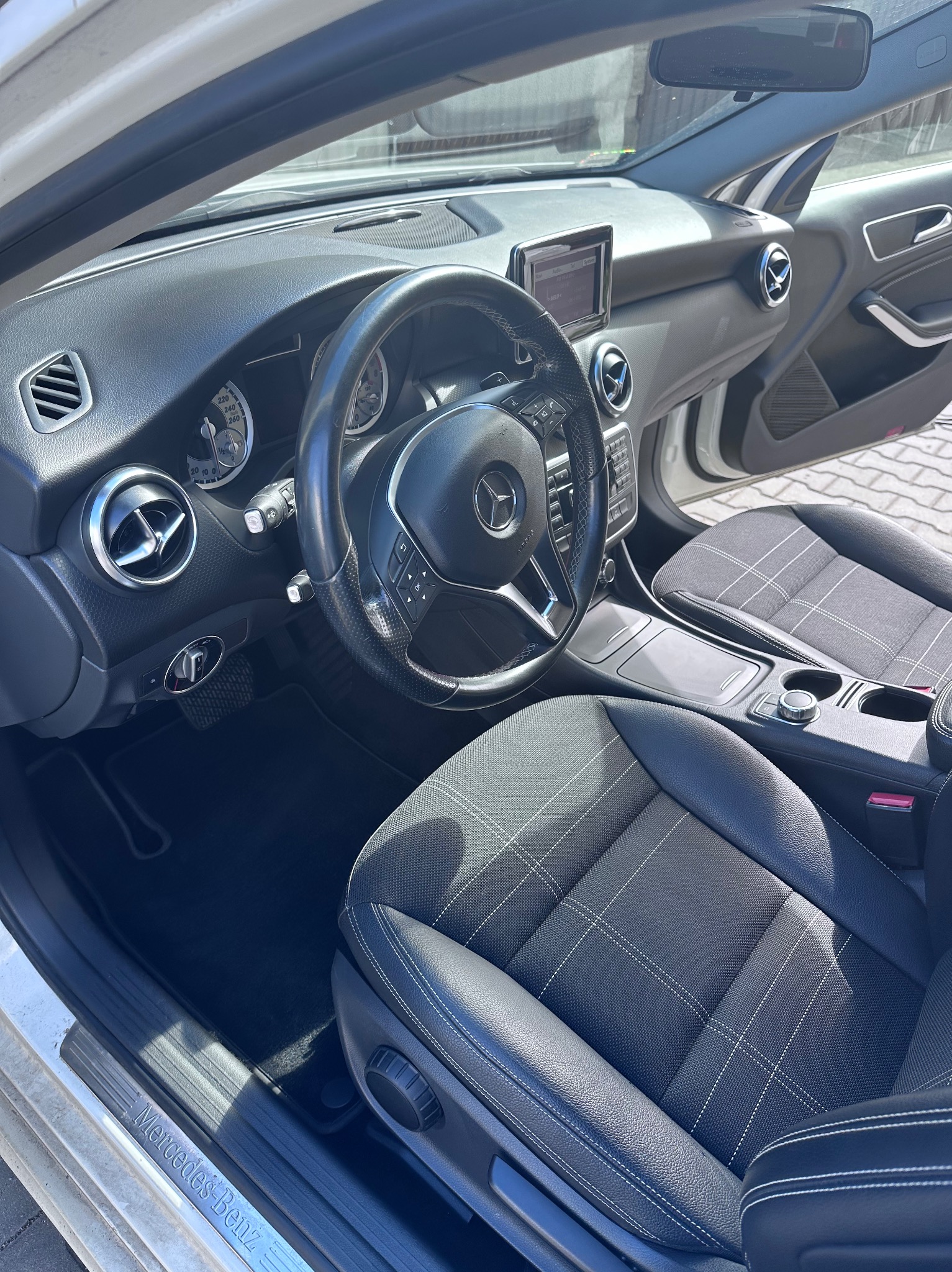 Wnętrze samochodu Mercedes-Benz klasy A z czarną deską rozdzielczą, kierownicą z logo, kratkami wentylacyjnymi i fotelami z ciemnej tkaniny w białą kratę.