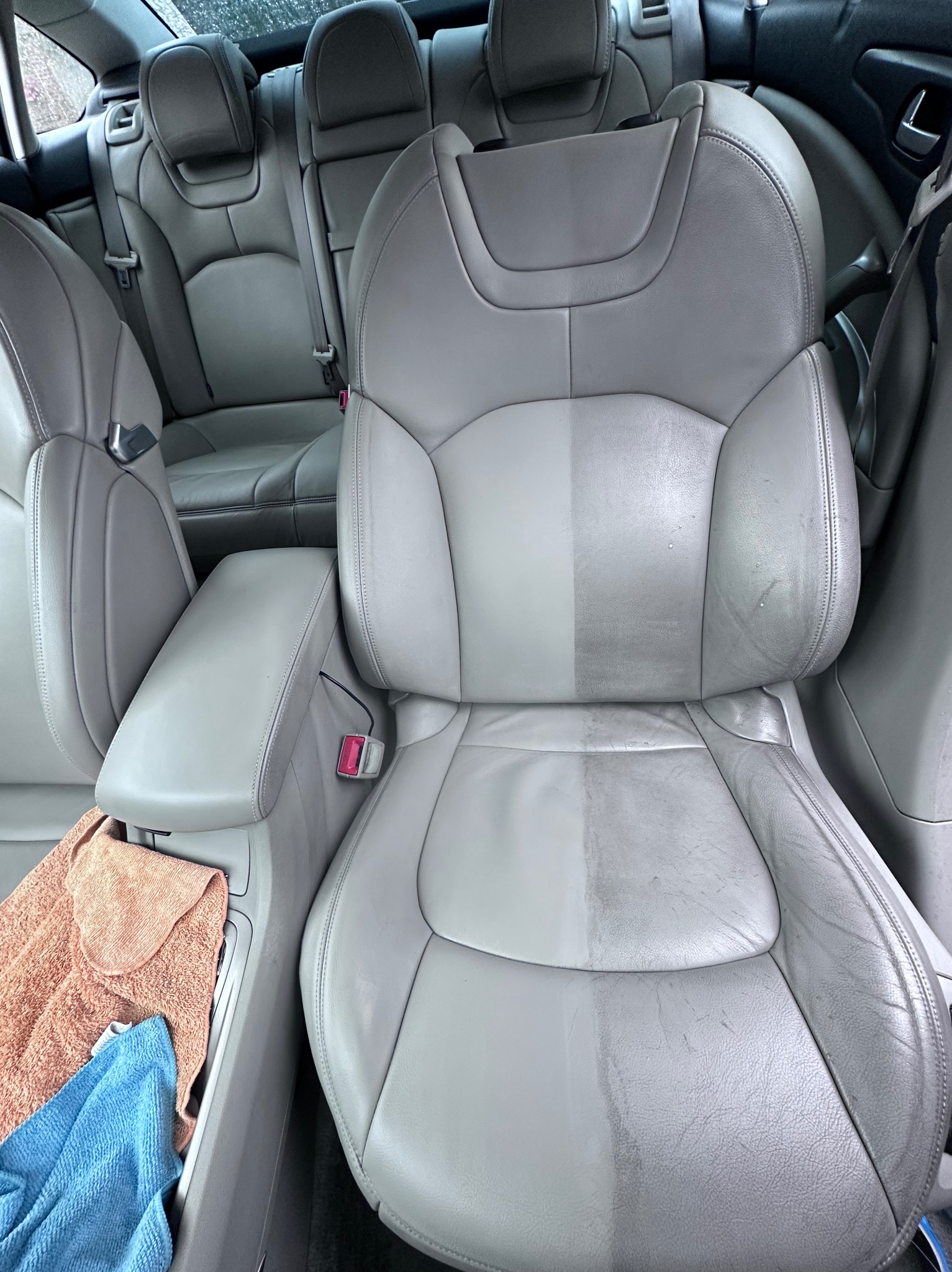 Szare skórzane siedzenie samochodowe w trakcie czyszczenia, widoczna wyraźna różnica między wyczyszczoną a brudną częścią, obok leżą złożone ręczniki z mikrofibry.