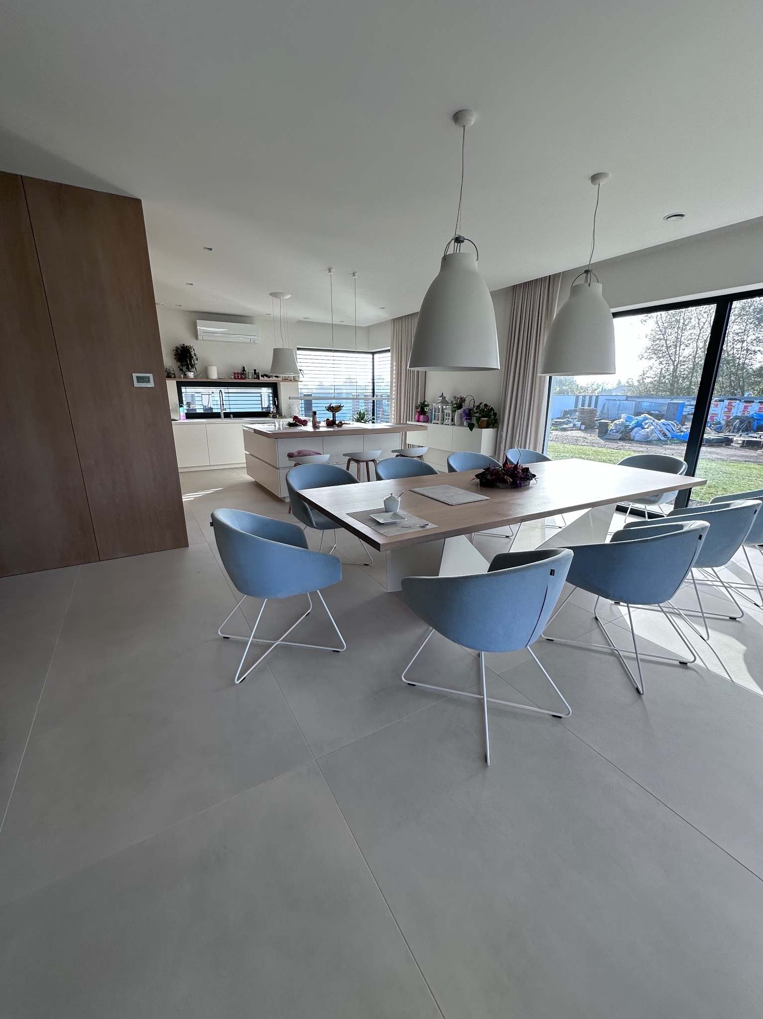 Jasne, przestronne wnętrze domu z otwartą kuchnią i jadalnią. W centrum duży, drewniany stół otoczony błękitnymi krzesłami na metalowych nogach. Nad stołem wiszą trzy białe lampy. Duże okno...