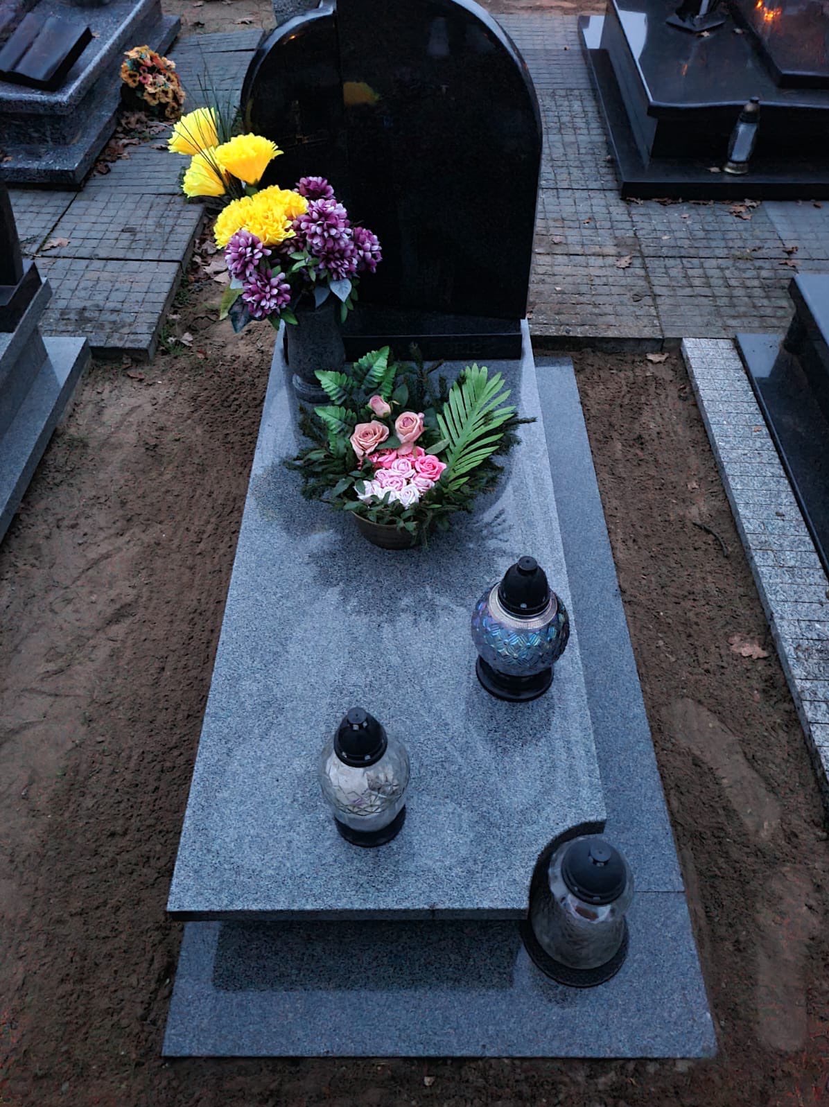 Granitowy nagrobek z kompozycjami kwiatowymi w odcieniach żółci, fioletu i różu, oraz trzema lampionami, umieszczony na cmentarzu.