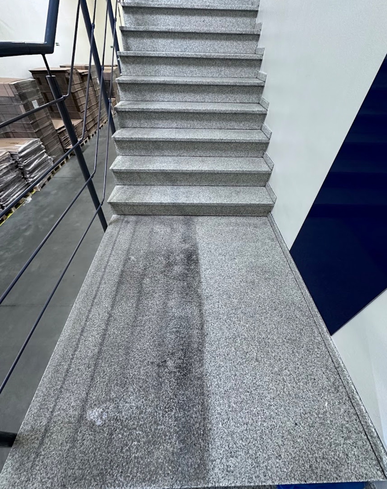 Schody z szarego granitu, widoczne ślady po czyszczeniu na podeście, w tle metalowa barierka i kartony.