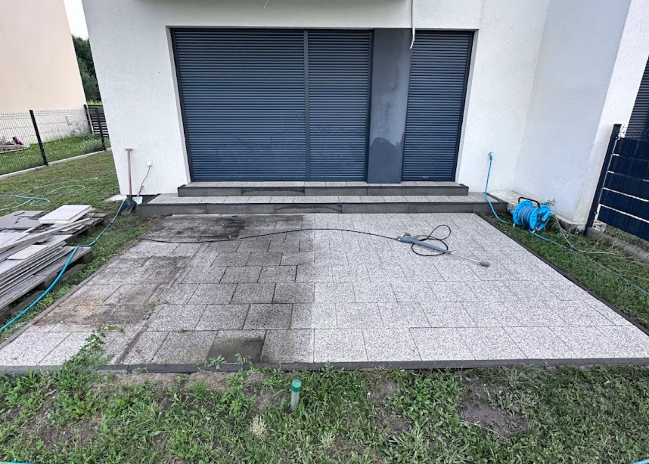 Czyszczenie kostki brukowej na tarasie domu jednorodzinnego, widoczna różnica między umytą a brudną powierzchnią, myjka ciśnieniowa w użyciu, paleta z kostką obok.