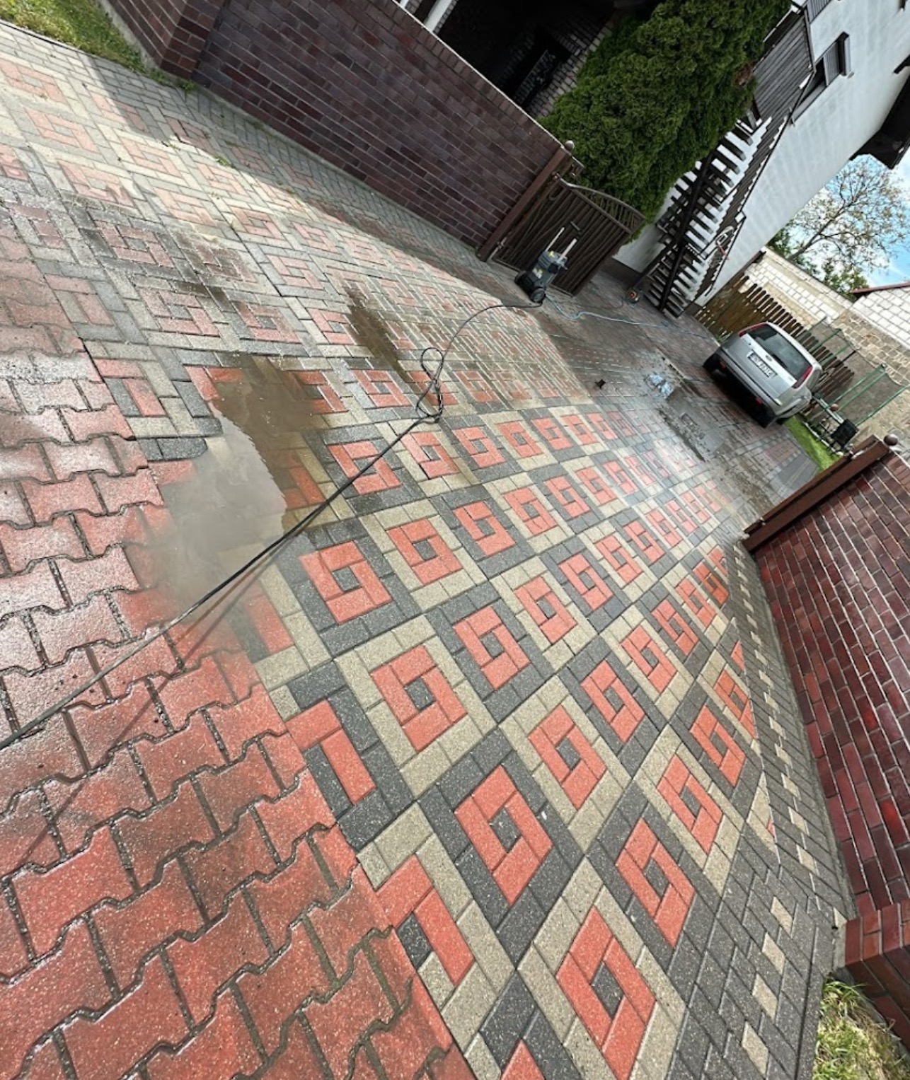 Częściowo umyta kostka brukowa z wzorem geometrycznym, widoczny fragment strumienia wody z myjki ciśnieniowej, w tle budynek mieszkalny i zaparkowany samochód.