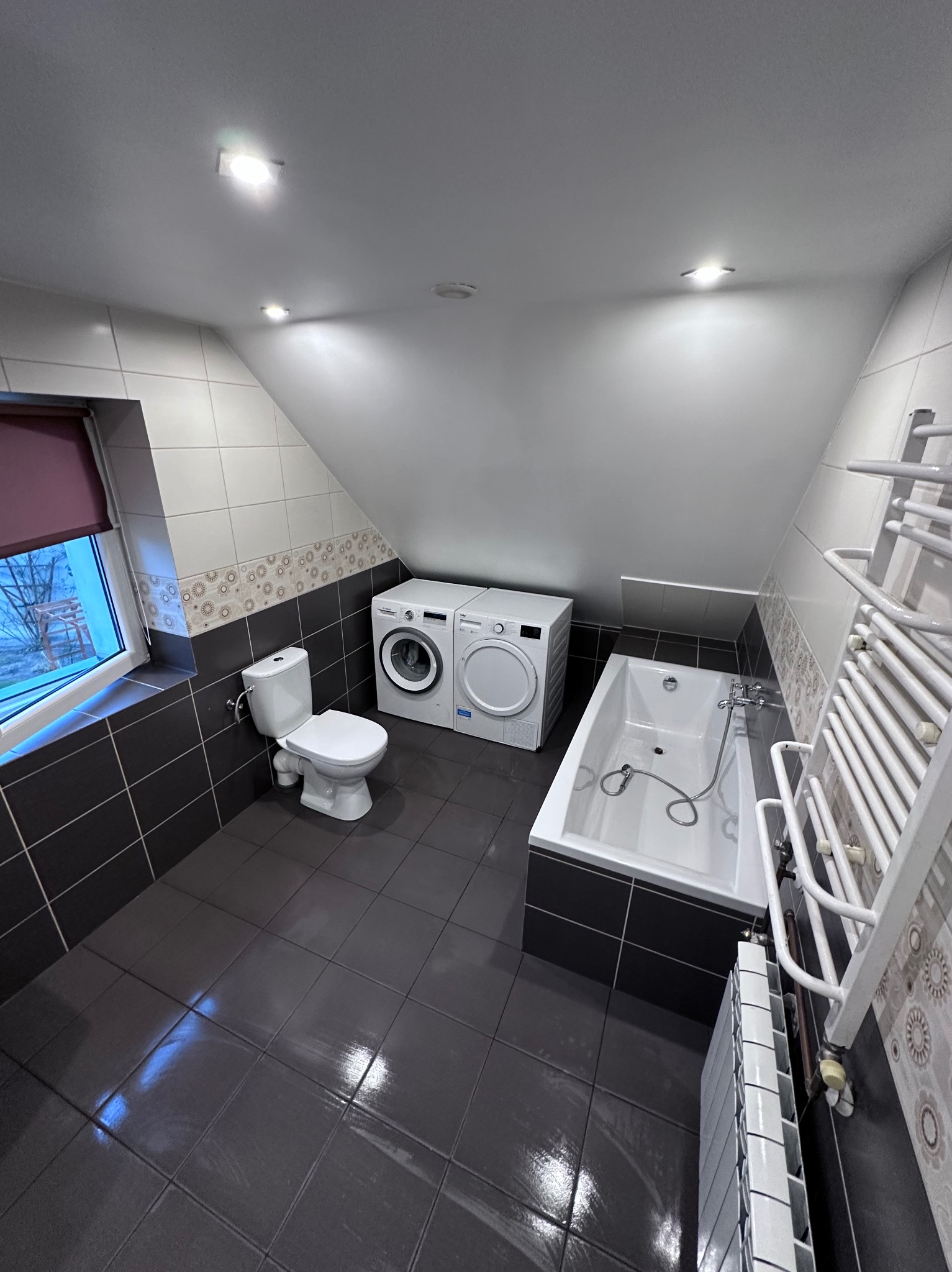 Wnętrze łazienki na poddaszu z pralką, suszarką, wanną i toaletą, wykończone ciemnymi płytkami z dekoracyjnym pasem.