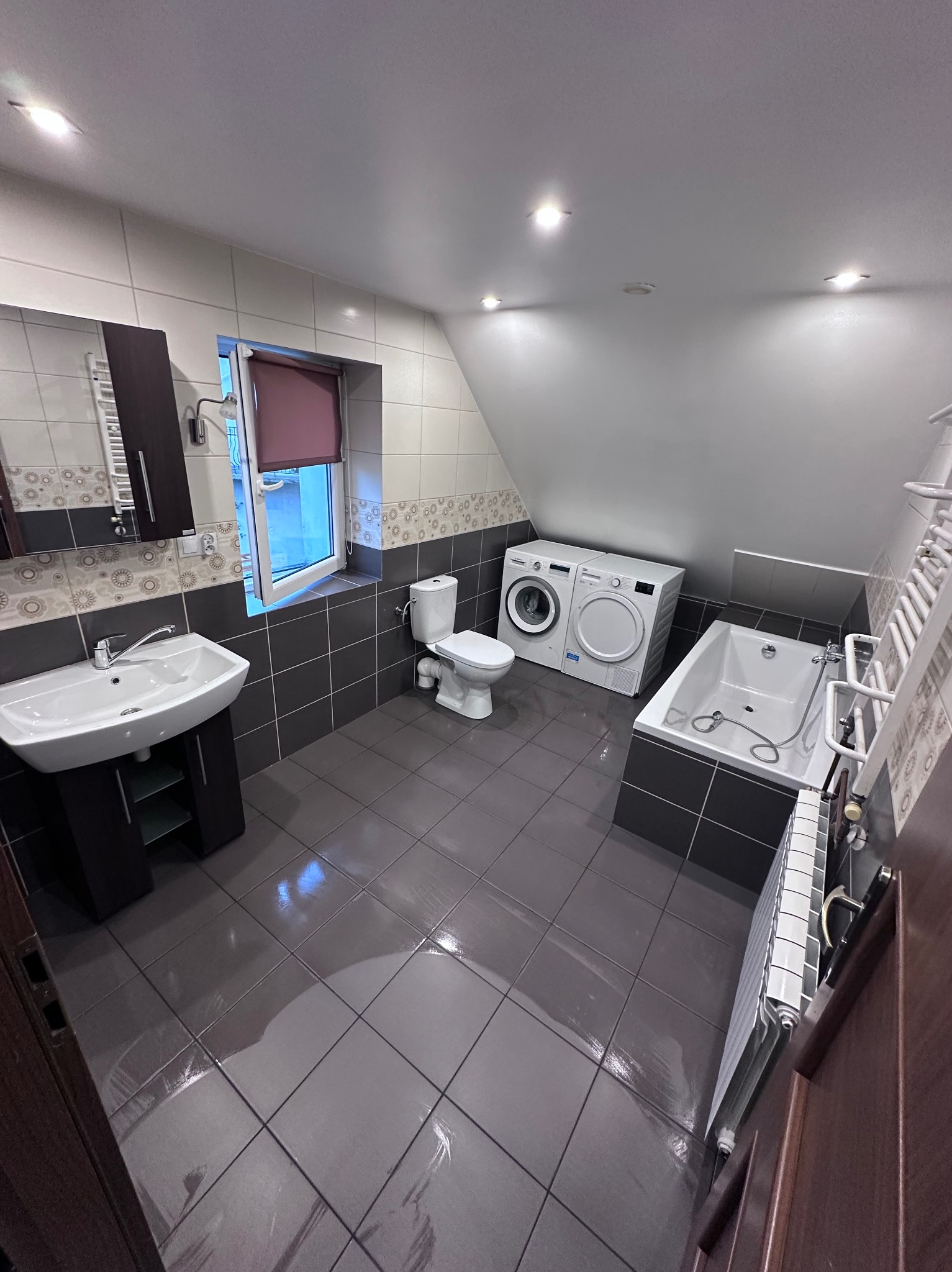 Wnętrze łazienki na poddaszu z umywalką, toaletą, wanną i pralko-suszarką, wykończone płytkami w odcieniach szarości i bieli.