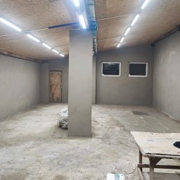 FIRMA BUDOWLANO-HANDLOWO-USŁUGOWA BAZ-BUD Mariusz BAZAN - Wnętrze pomieszczenia po nałożeniu tynku cementowo-wapiennego na ściany i filar, widoczne dwa okna, drzwi z płyty OSB, instalacja pod sufitem i worki z materiałami budowlanymi.