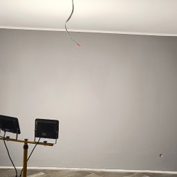 FIRMA BUDOWLANO-HANDLOWO-USŁUGOWA BAZ-BUD Mariusz BAZAN - Pokój po malowaniu na szaro, widoczne przewody elektryczne, lampy halogenowe na statywie i fragment podłogi