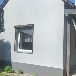 FIRMA BUDOWLANO-HANDLOWO-USŁUGOWA BAZ-BUD Mariusz BAZAN - Otynkowana elewacja domu jednorodzinnego w odcieniach szarości, z widocznym fragmentem dachu z dachówką, oknem z roletą i elementami ogrodu.