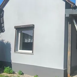 Otynkowana elewacja domu jednorodzinnego w odcieniach szarości, z widocznym fragmentem dachu z dachówką, oknem z roletą i elementami ogrodu.