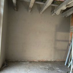 FIRMA BUDOWLANO-HANDLOWO-USŁUGOWA BAZ-BUD Mariusz BAZAN - Świeżo nałożony tynk maszynowy na ścianie w pomieszczeniu w trakcie remontu, widoczne zabezpieczone elementy konstrukcyjne sufitu i resztki materiałów budowlanych na podłodze.