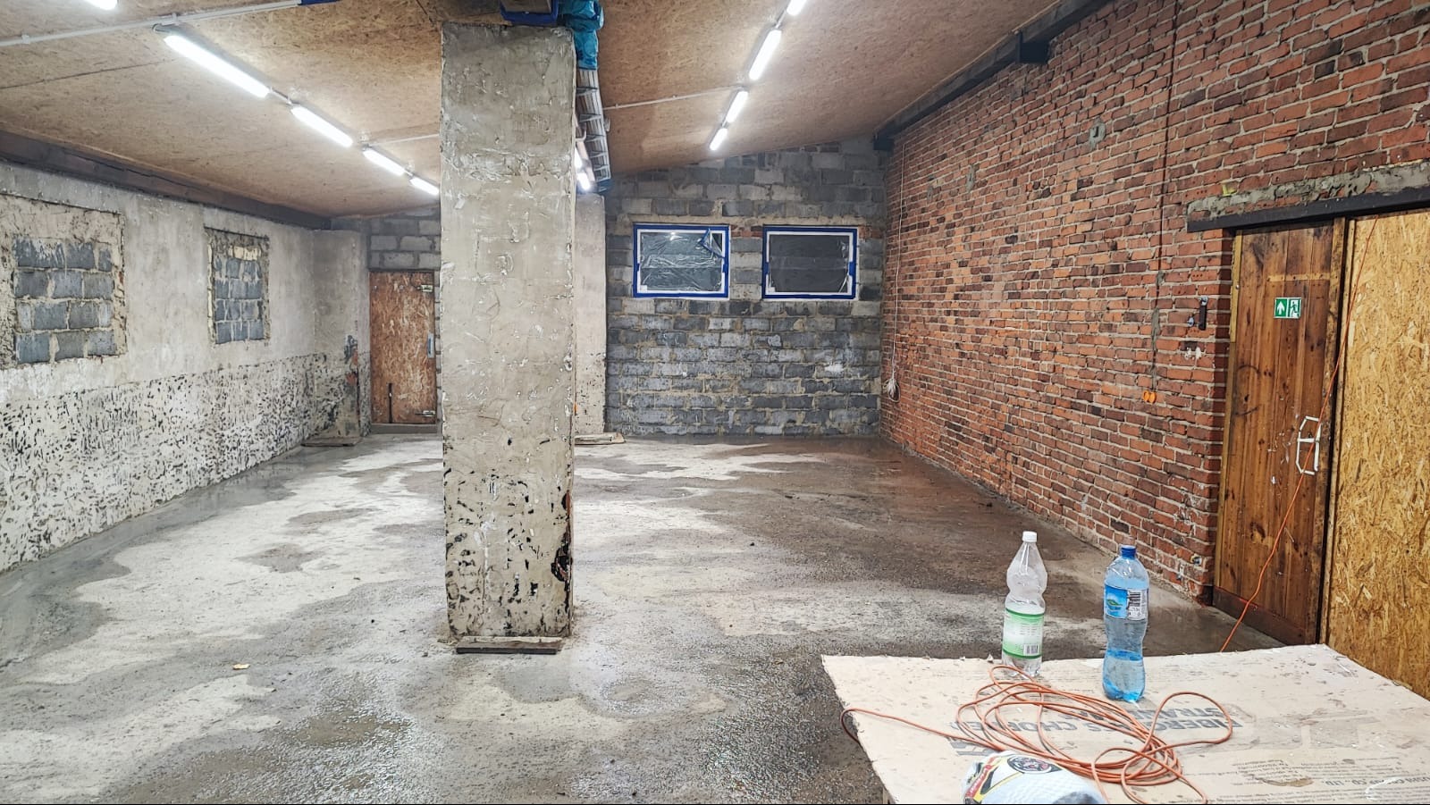 Wnętrze w trakcie remontu: surowa ceglana ściana, betonowa podłoga z widocznymi śladami wilgoci, zabezpieczone okna folią malarską, kolumna z resztkami tynku, widoczne butelki z wodą i kabel...